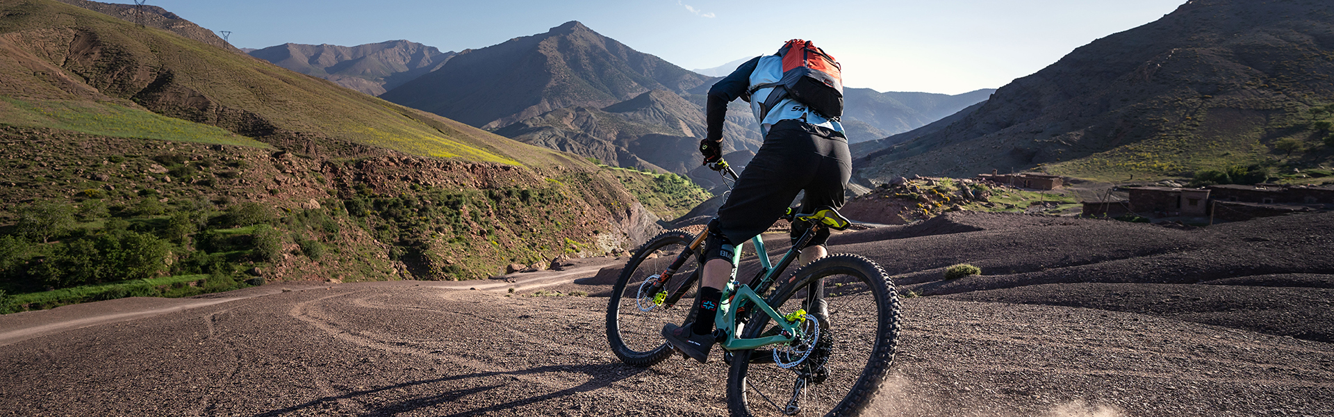 Un ciclista di mountain bike solleva una nuvola di polvere su un sentiero roccioso, sullo sfondo di un vasto paesaggio montano sotto un cielo azzurro e limpido.