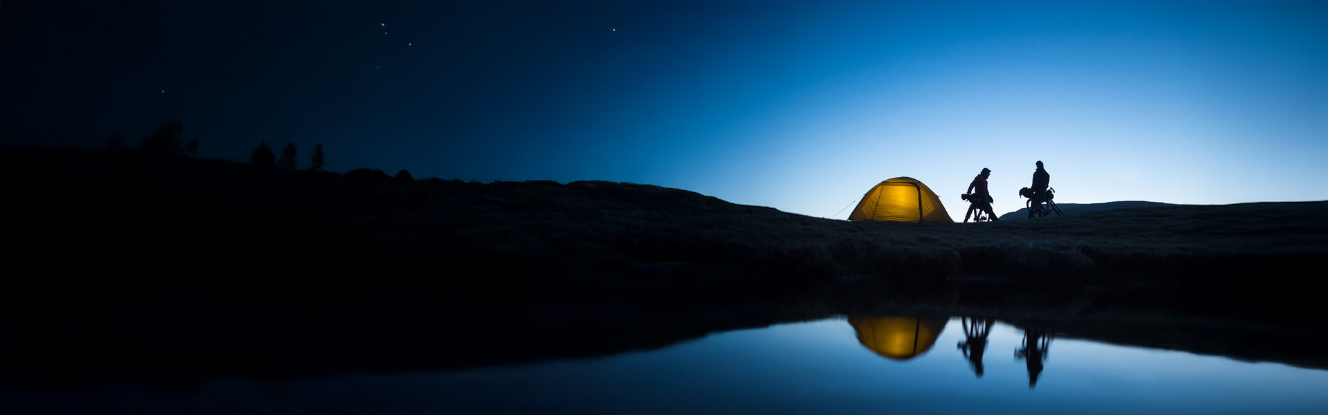Silhouettes of two people and a glowing tent under a starry sky, reflected in a calm body of water at dusk.