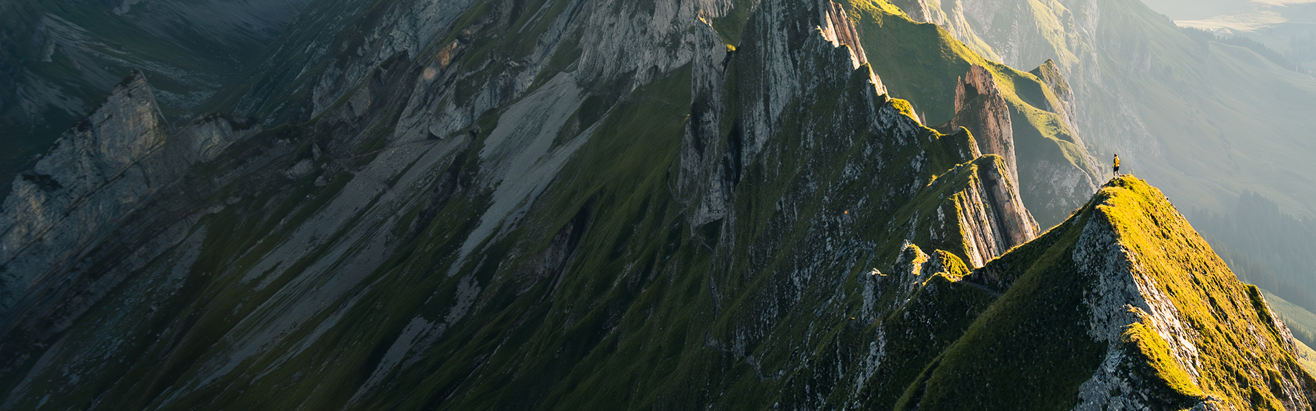 Bergrücken im flachen Abendlicht mit scharfen Gipfeln, bedeckt mit Gras und Felsen, unter einem klaren Himmel, mit Panoramablick auf ferne Täler.