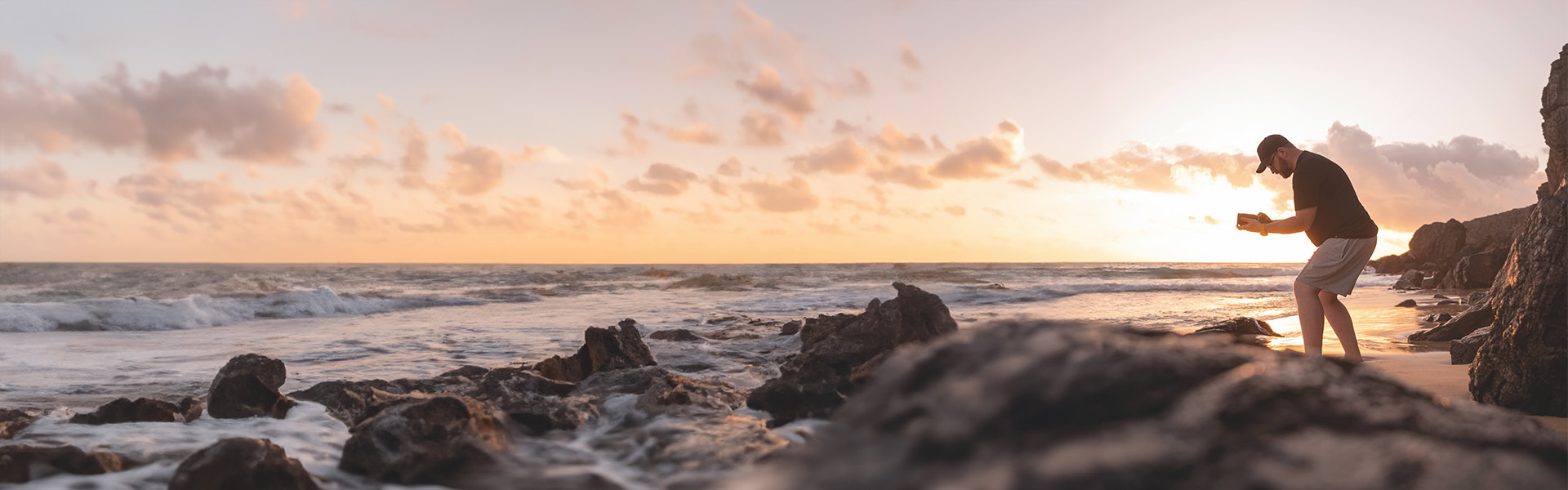 Una persona è in piedi su una spiaggia rocciosa al tramonto, mentre scatta foto con una fotocamera. Il cielo è coperto di nuvole e le onde si infrangono sulla riva.