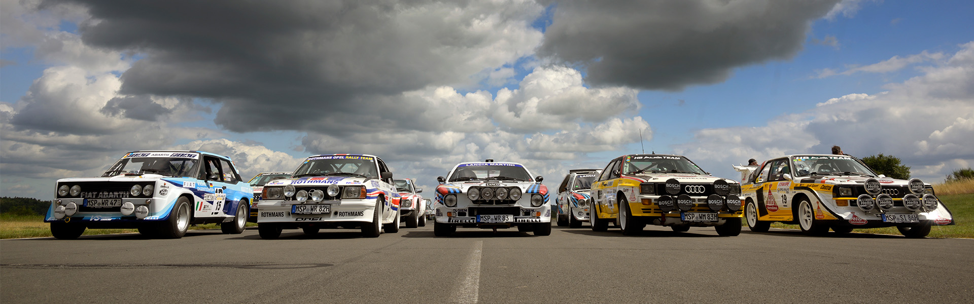 Five vintage rally cars lined up on a road under a cloudy sky, each with distinctive decals and front-mounted lights.