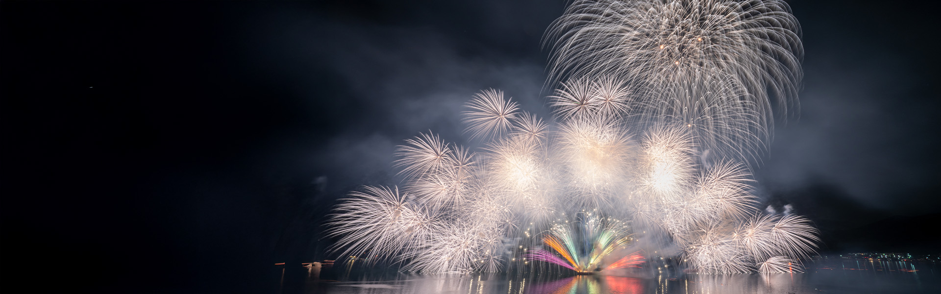 Ein lebendiges Feuerwerk über dem Wasser, mit weißen und bunt leuchtenden Explosionen vor einem dunklen Nachthimmel. Die Reflexionen der Lichtbursts spiegeln sich auf der Wasseroberfläche.