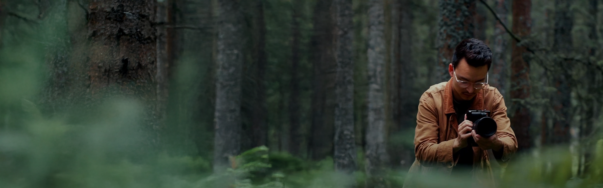 A person in a brown jacket is taking a photo with a camera in a dense, green forest.