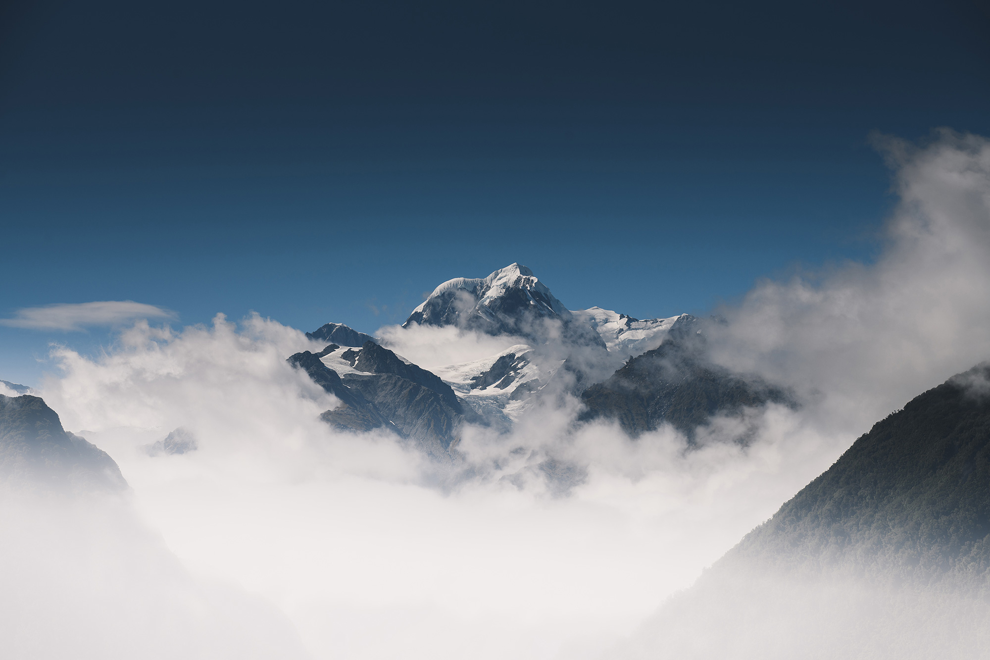 Ein schneebedeckter Berggipfel, umgeben von Wolken, unter einem strahlend blauen Himmel, mit dunklen Berghängen im Vordergrund.