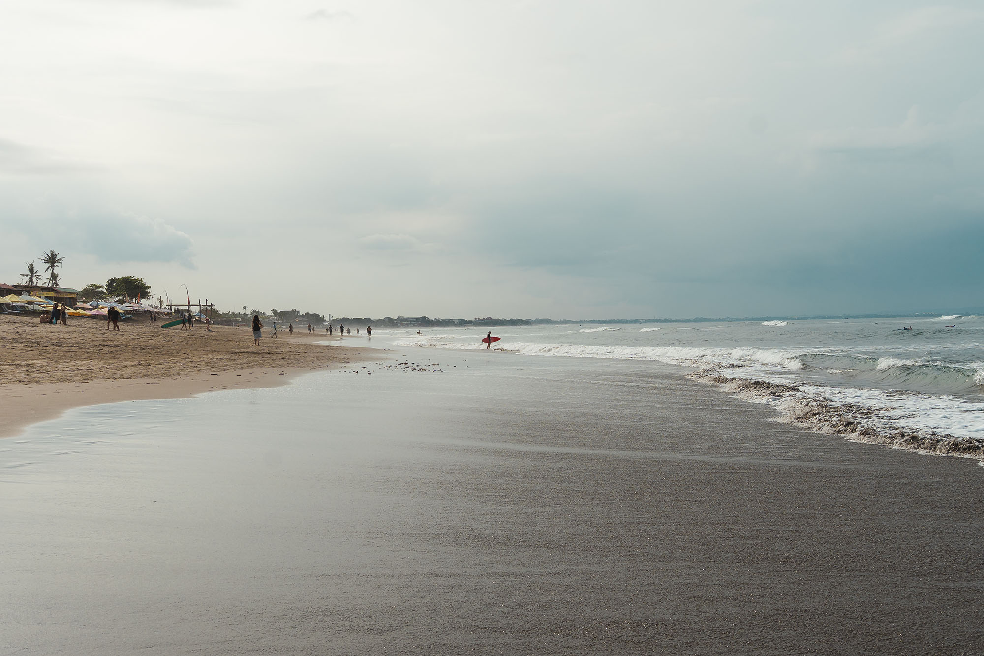 Eine wolkige Strandszene mit sanften Wellen, einigen Spaziergängern und einem Surfer, der ein rotes Surfbrett am Ufer entlangträgt.