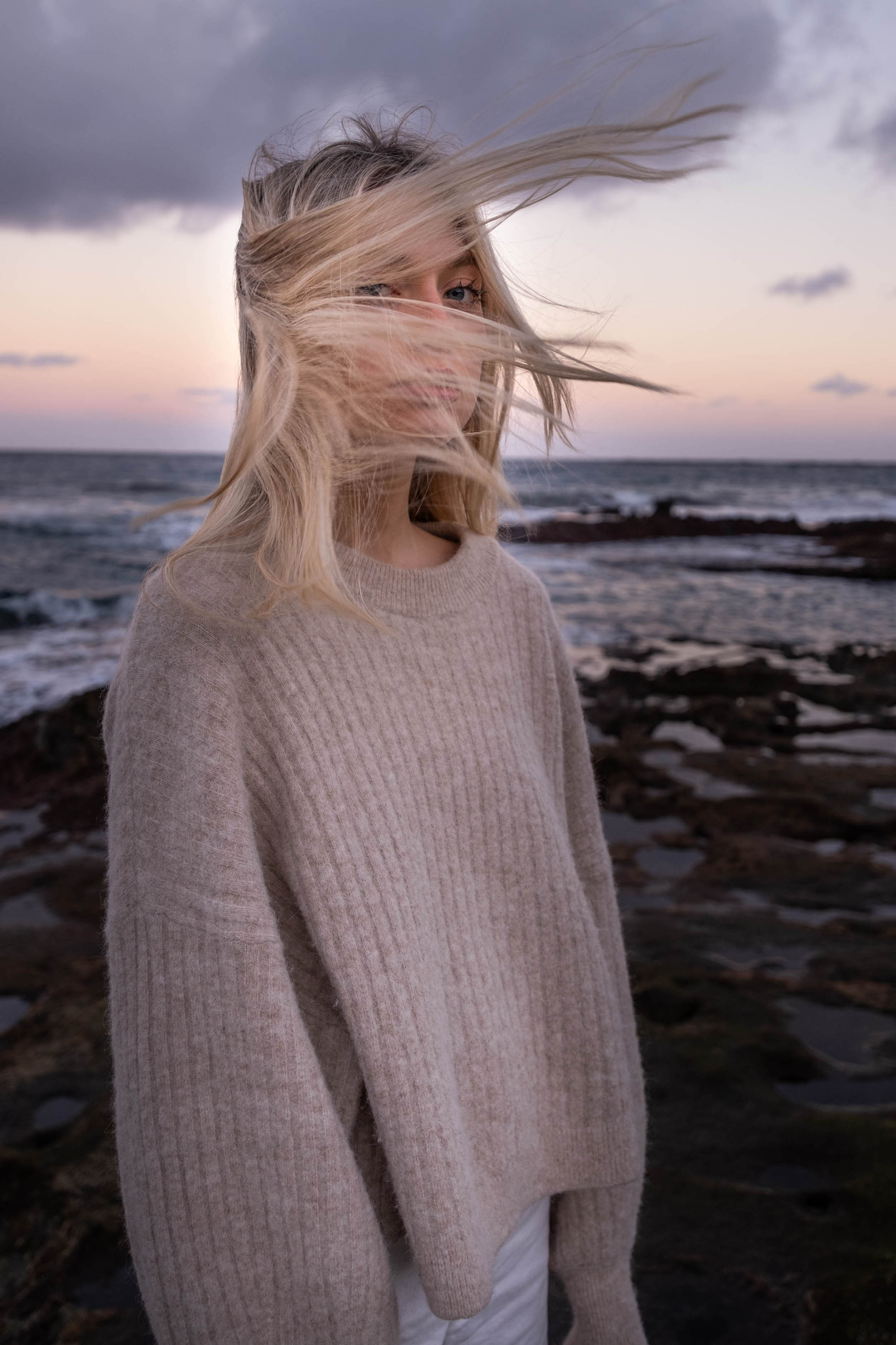 Eine Frau in einem beigen Pullover steht bei Sonnenuntergang am Meer, während ihr der Wind die Haare ins Gesicht weht. Im Hintergrund sieht man dunkle Felsen und Wellen.