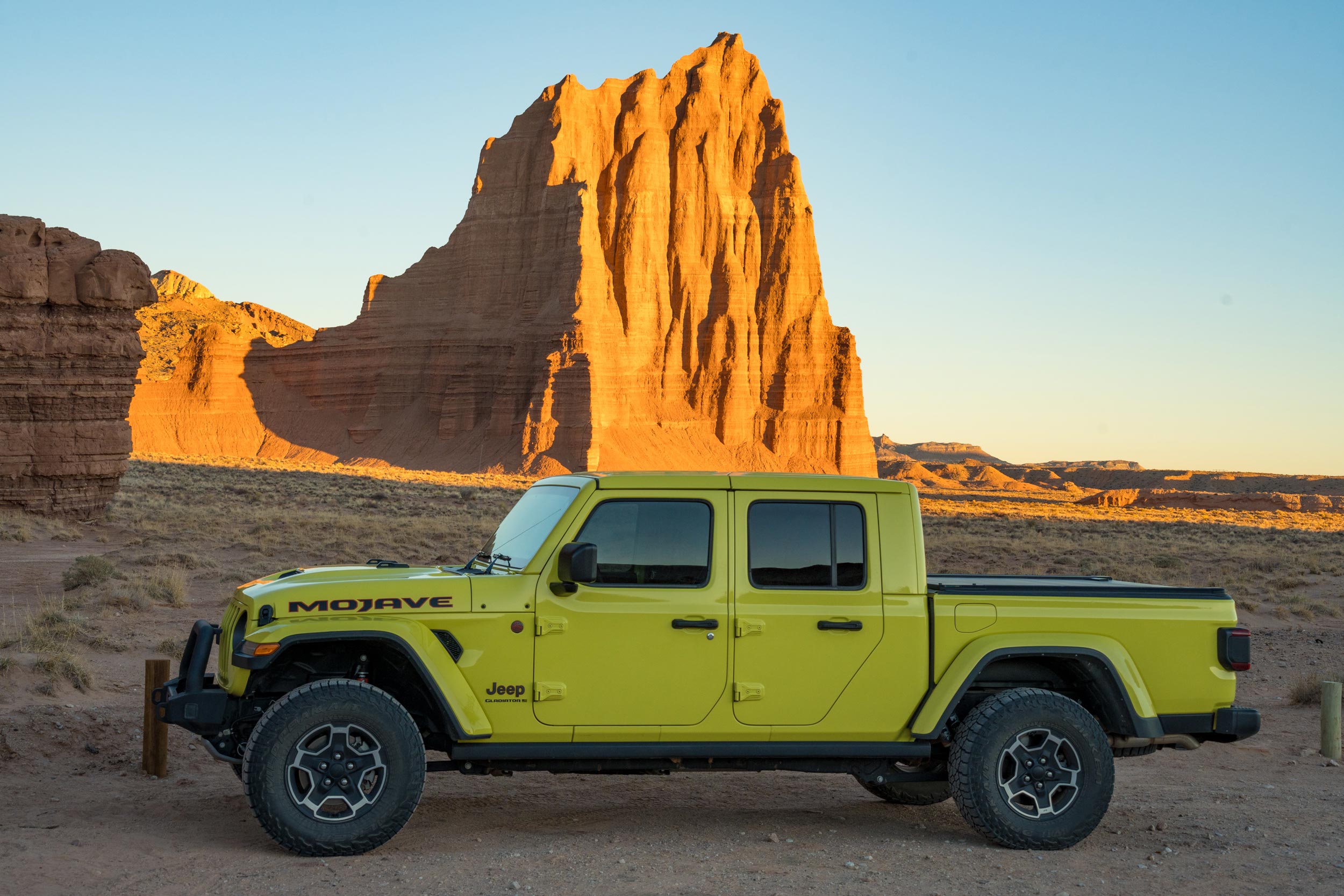 Yellow Jeep Gladiator parked in a desert landscape with a towering red rock formation under a clear blue sky at sunset.