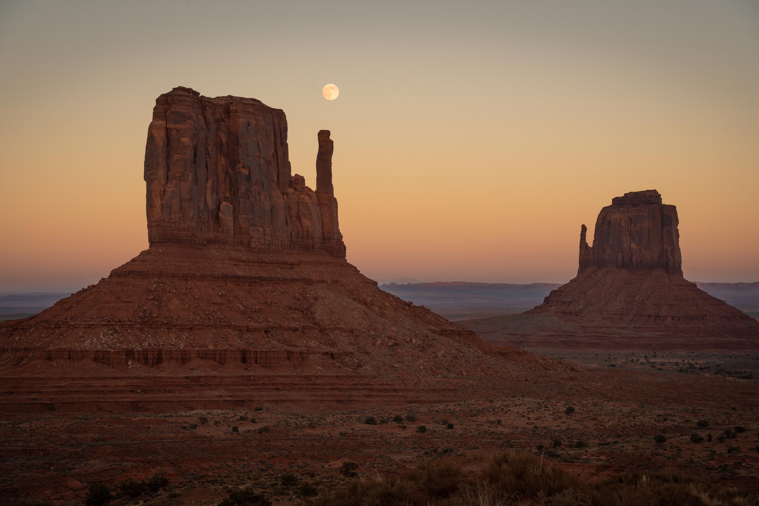 Monument Valley landscape at dusk with a full moon rising above two iconic sandstone buttes under a subtle gradient sky.