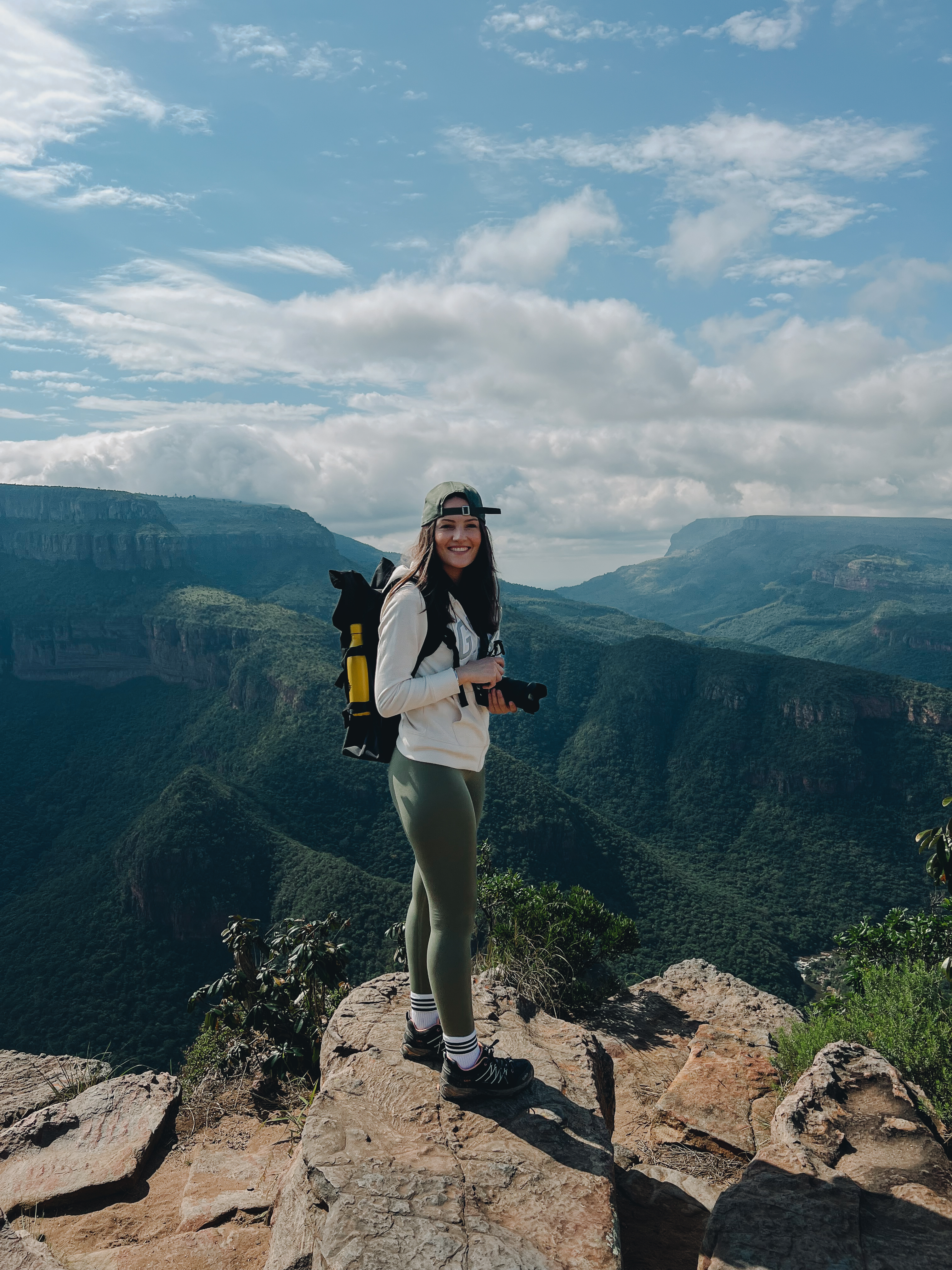 Una donna in abbigliamento da trekking si trova su una scogliera rocciosa, con una macchina fotografica in mano. Sullo sfondo si apre un’ampia distesa di montagne verdi sotto un cielo nuvoloso.