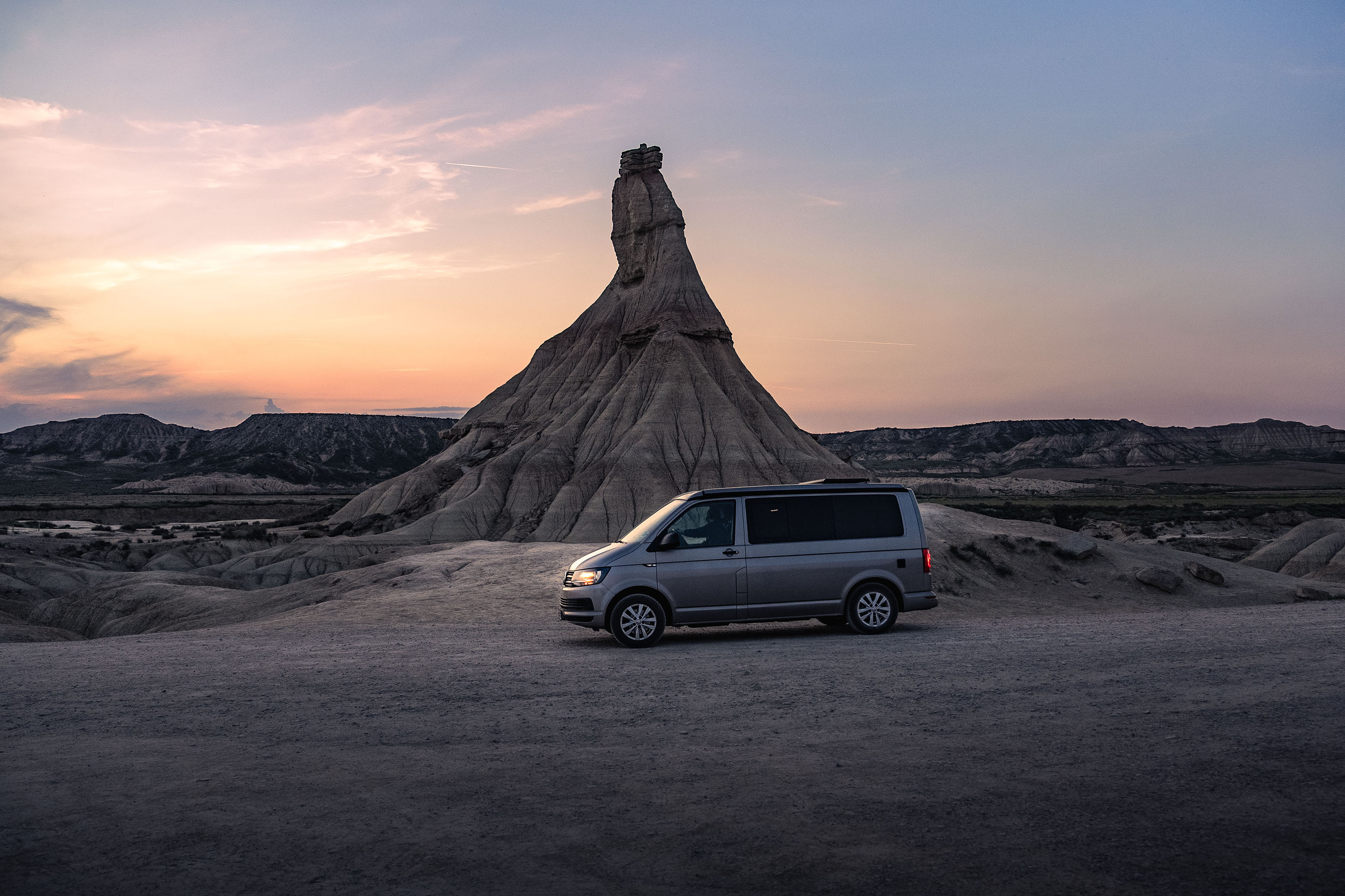 Un furgone argentato parcheggiato davanti a una formazione rocciosa unica al tramonto, con un cielo colorato sullo sfondo.