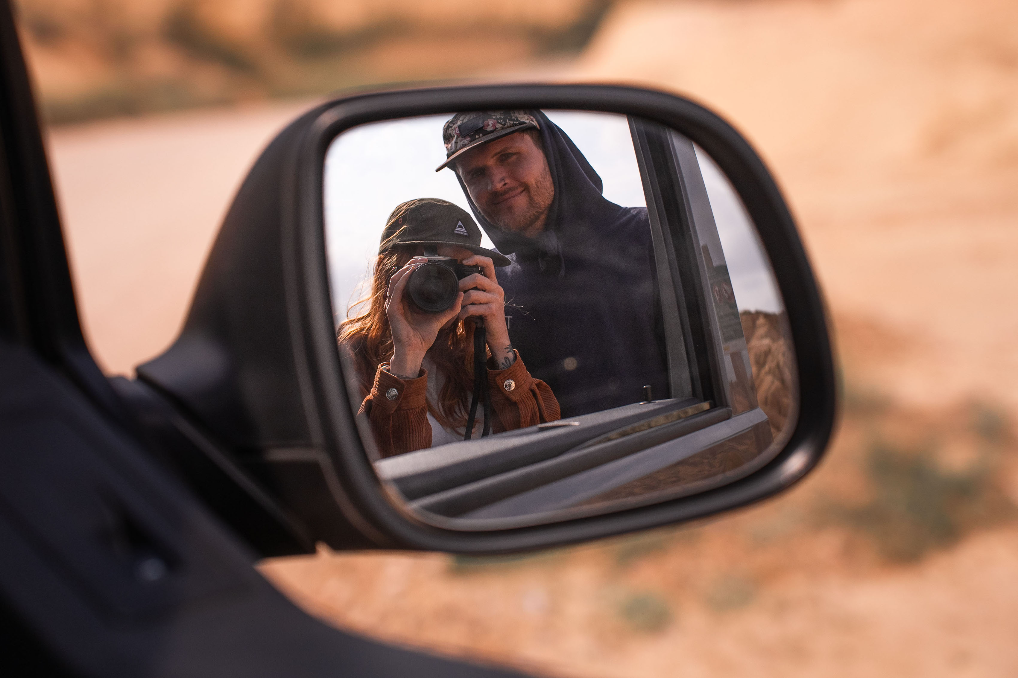 Riflesso di due persone nello specchietto laterale di un’auto, una delle quali tiene in mano una fotocamera, con un paesaggio desertico sullo sfondo.