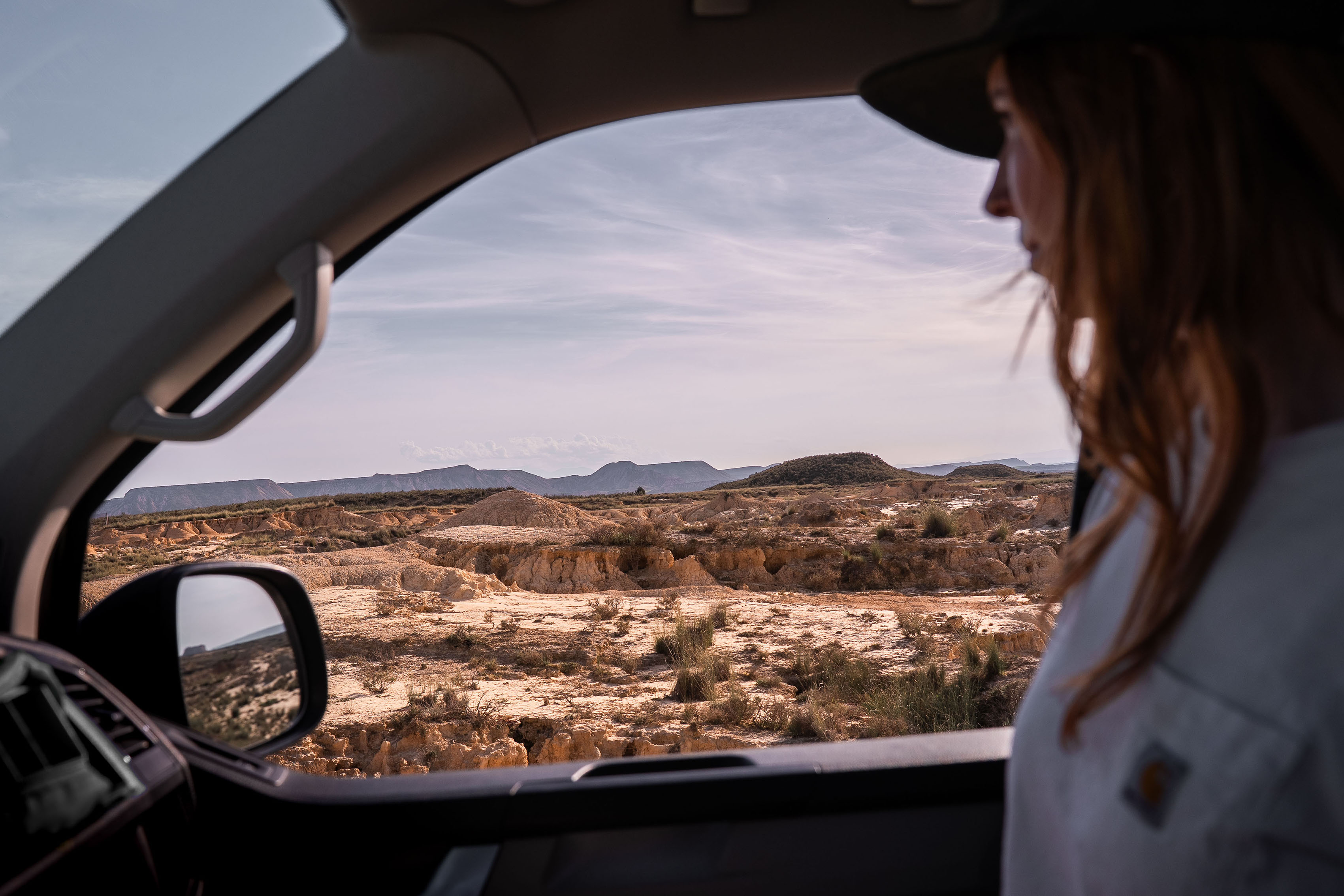 Una persona in auto osserva, attraverso il finestrino, un vasto paesaggio desertico e arido con colline lontane sotto un cielo limpido.