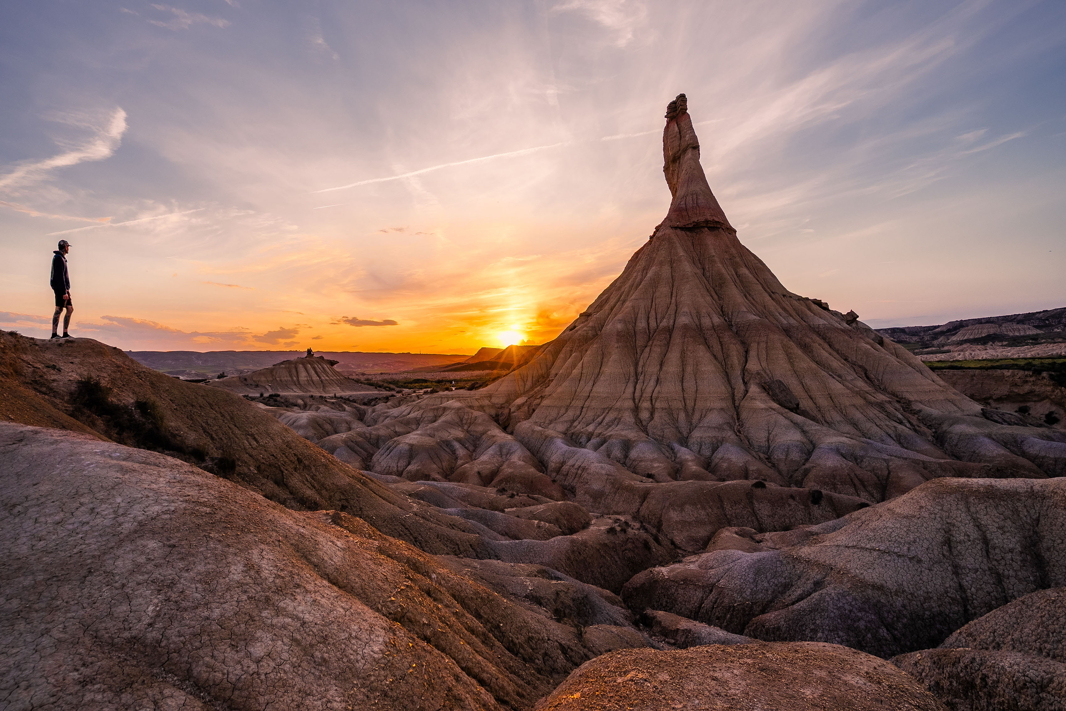 Una persona sta su un terreno roccioso, osservando il tramonto dietro una formazione rocciosa unica e appuntita, in un paesaggio desertico.