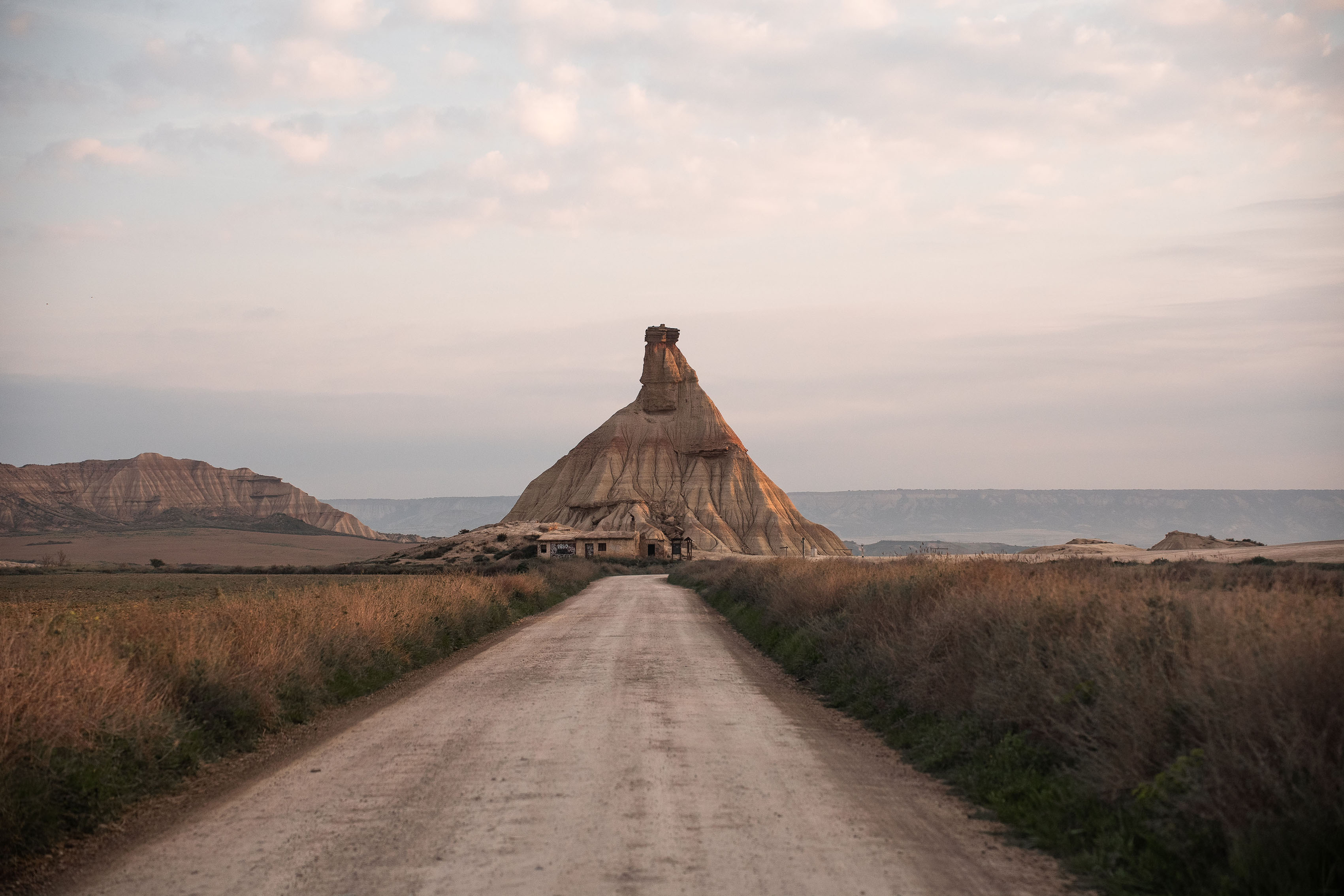 Una strada sterrata conduce a una formazione rocciosa unica a forma di cono, sotto un cielo nuvoloso e circondata da un terreno pianeggiante e erboso.