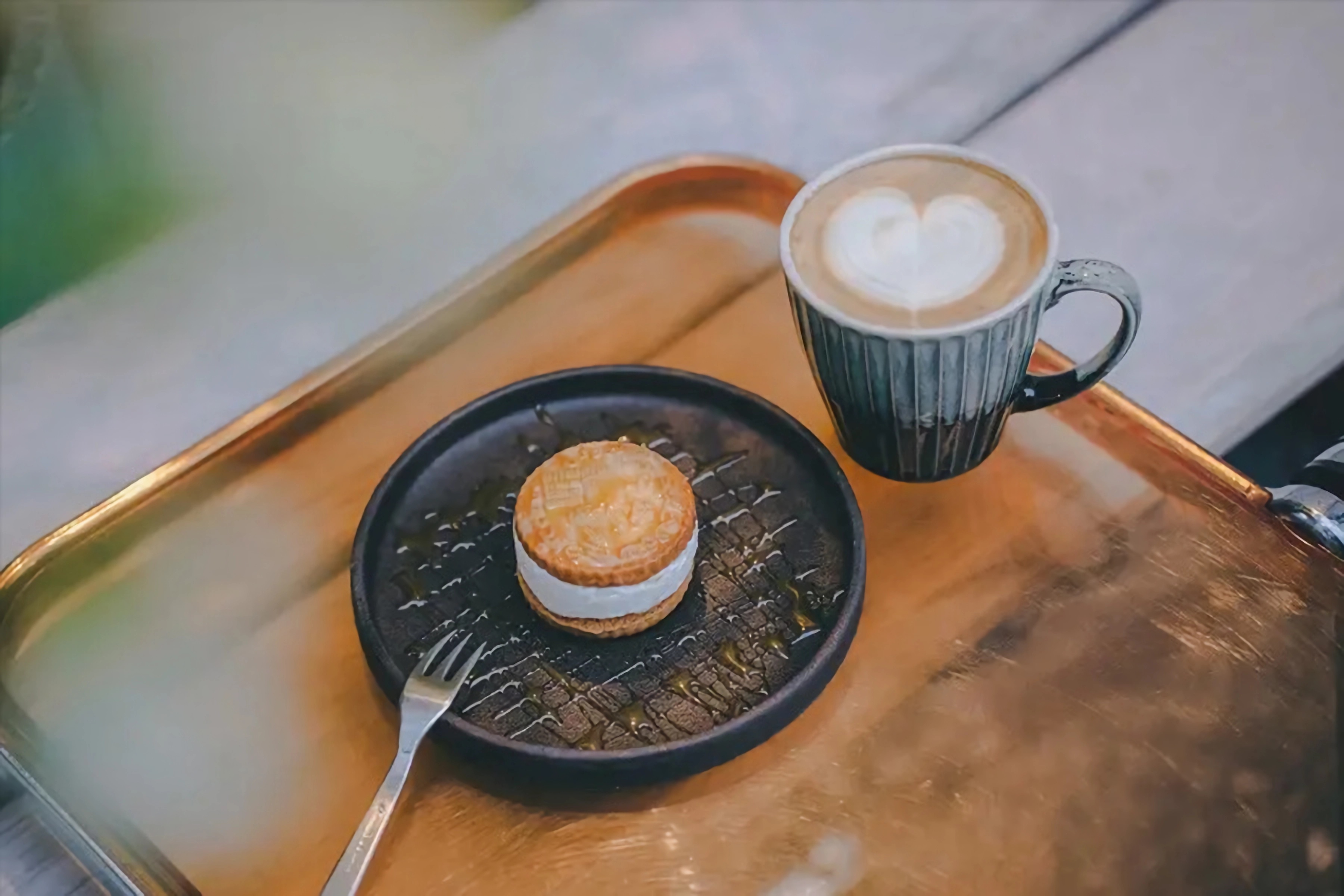 Ein Holztisch mit einem Tablett, darauf eine Tasse Kaffee und ein kleiner Kuchen auf einem Teller, vor einem unscharfen, grünen Naturhintergrund.