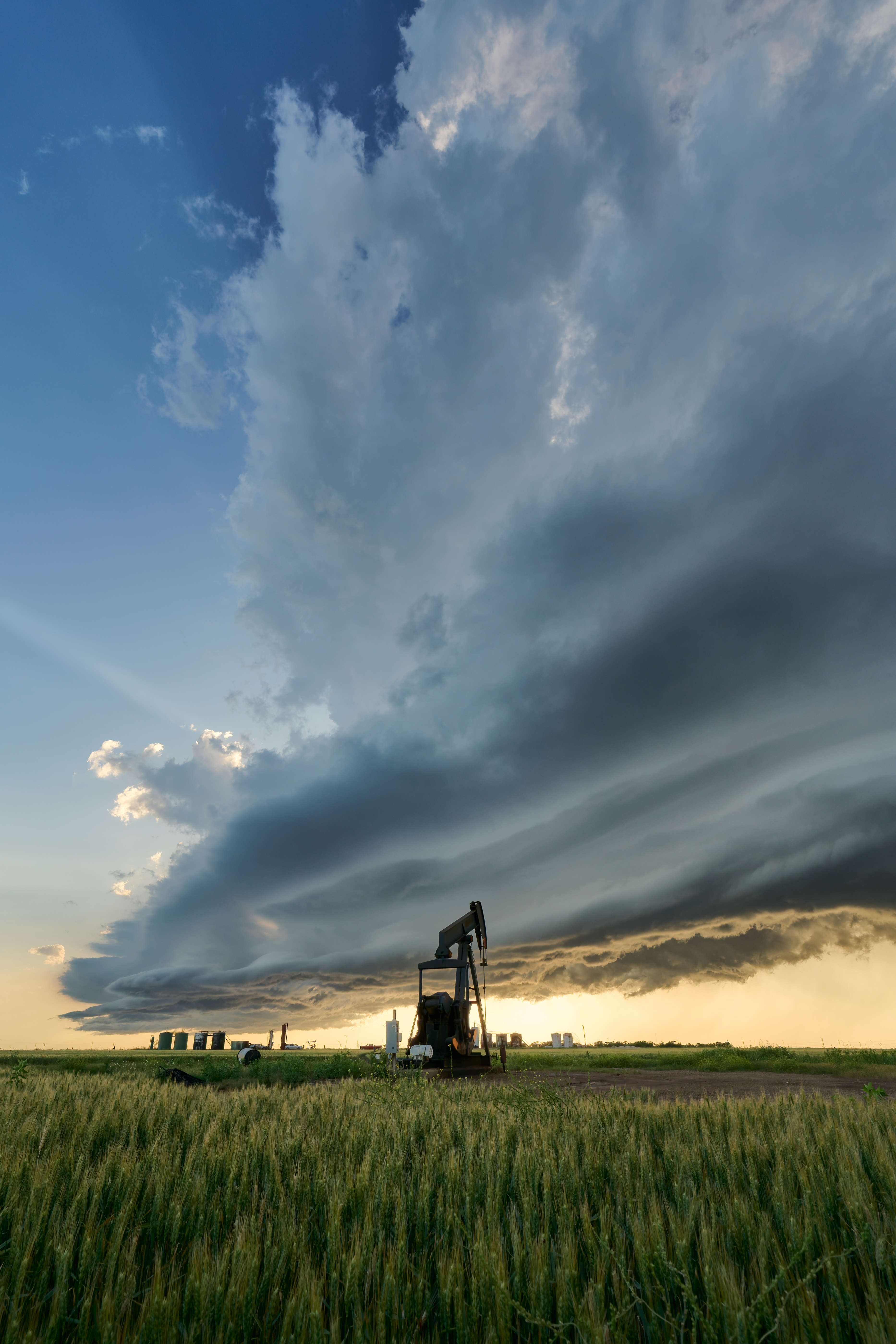 Dramatic storm clouds loom over an oil pumpjack in a vast green field, with a distant horizon and a hint of sunlight peeking through.