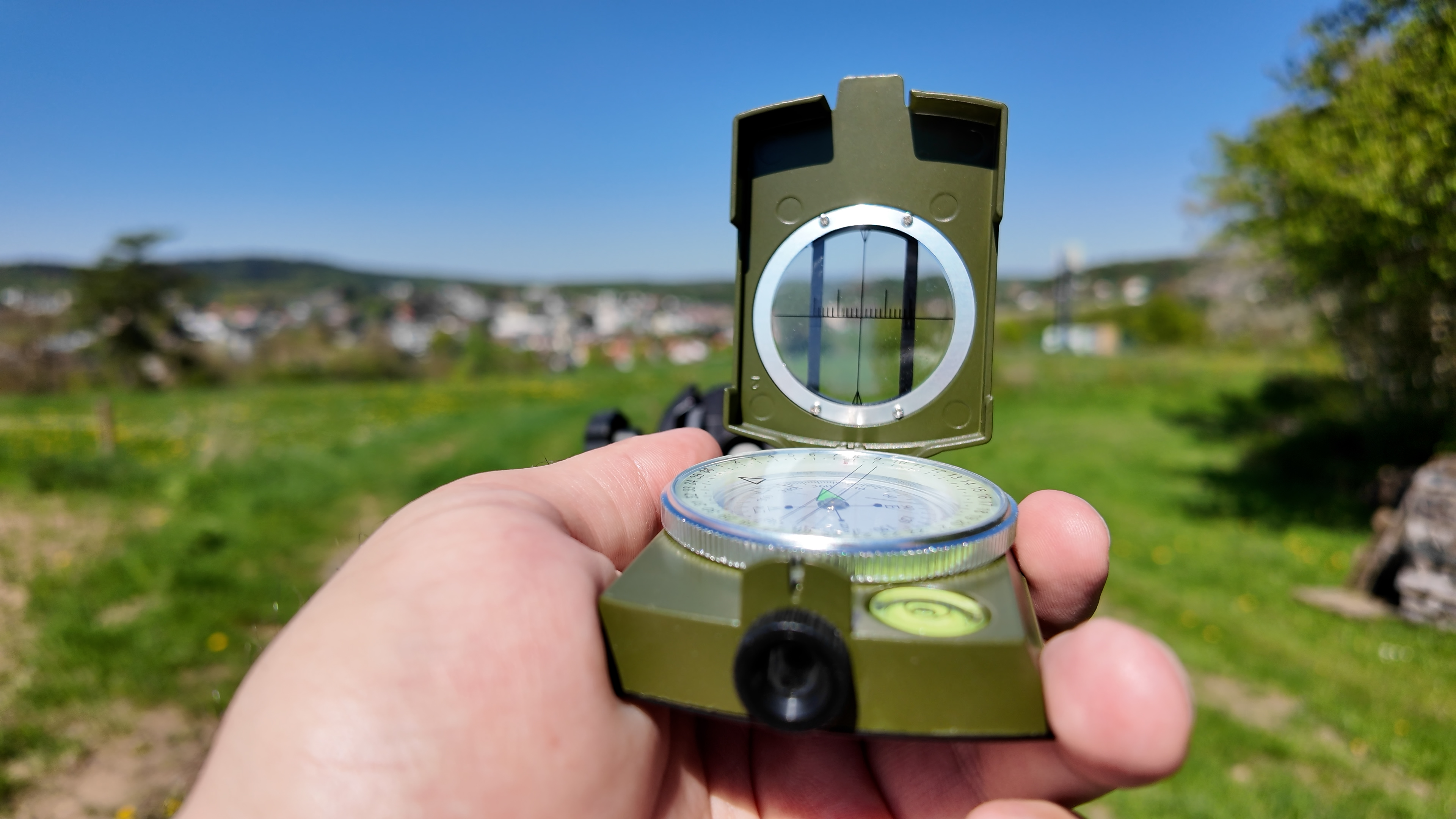 In the foreground, a hand is holding a green compass; in the background, a blurred landscape can be seen beneath a bright blue sky.