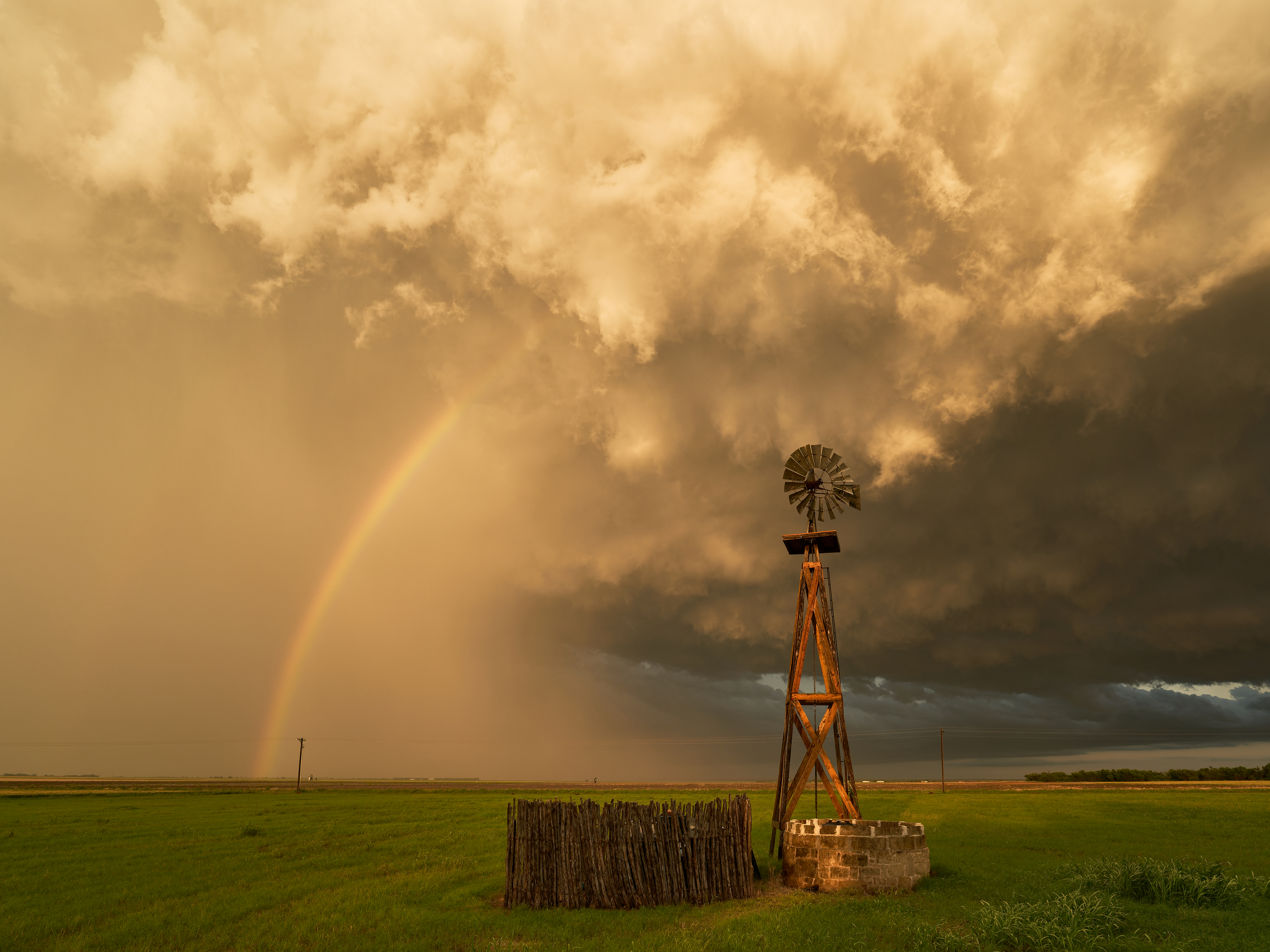 A dramatic sky with a rainbow and dark clouds over a green field, featuring an old windmill and wooden fence in the foreground.
