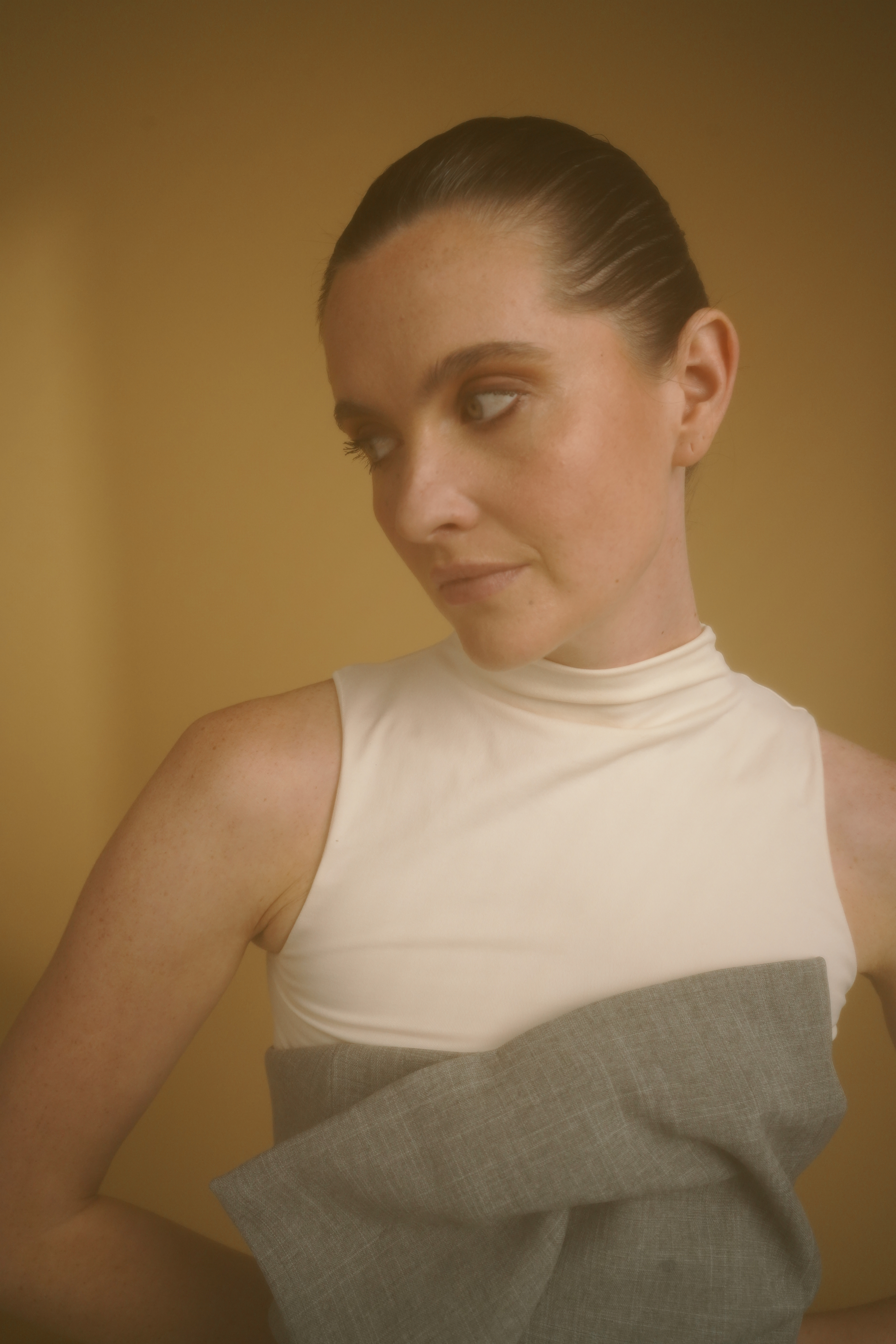 Een vrouw met naar achter gekamd haar draagt een mouwloos topje en kijkt opzij, tegen een zachte achtergrond in warme tinten.