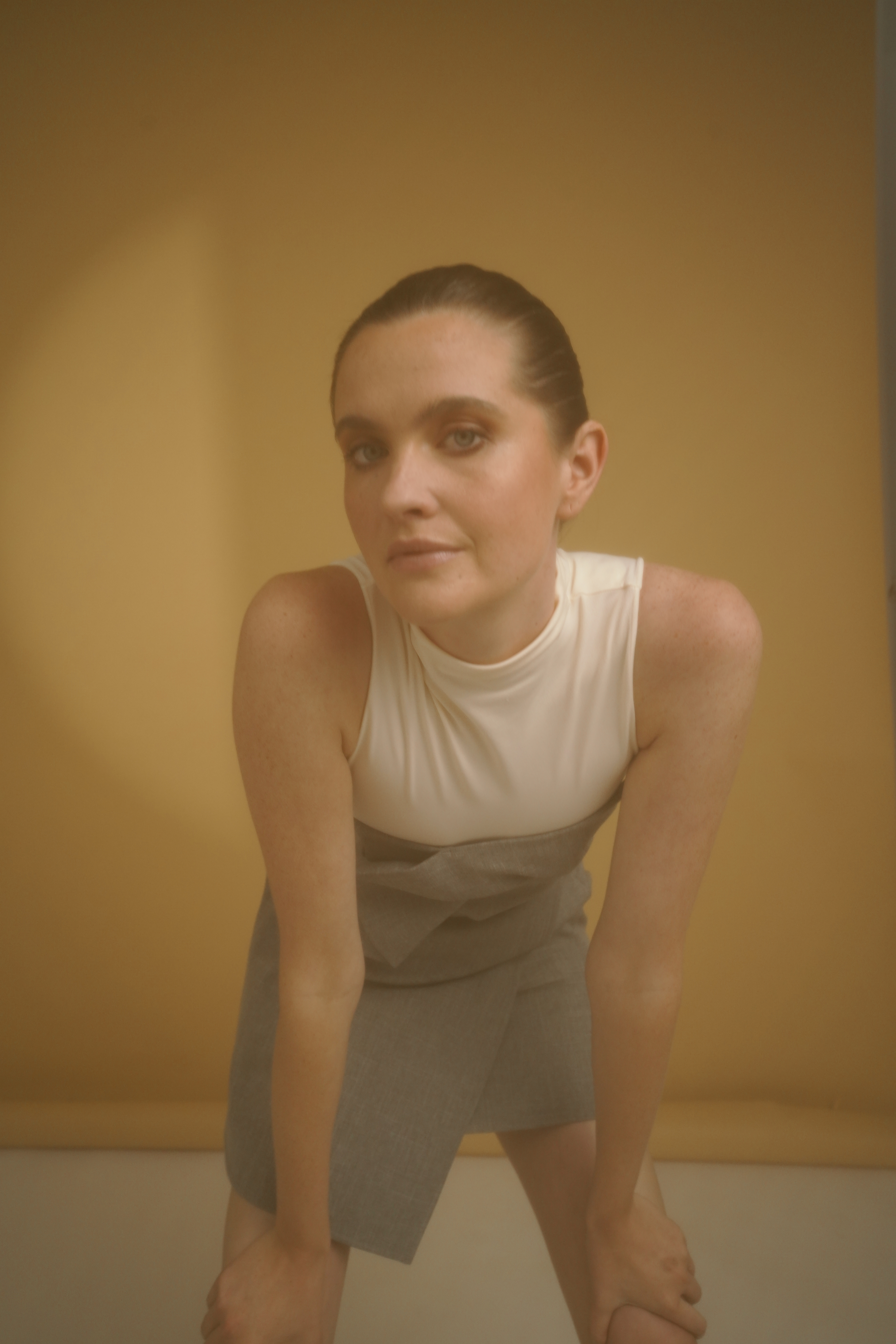 Een vrouw met een wit mouwloos topje en een grijze rok poseert voor een warme beige achtergrond. Ze leunt naar voren en kijkt met een zelfverzekerde uitdrukking in de camera.