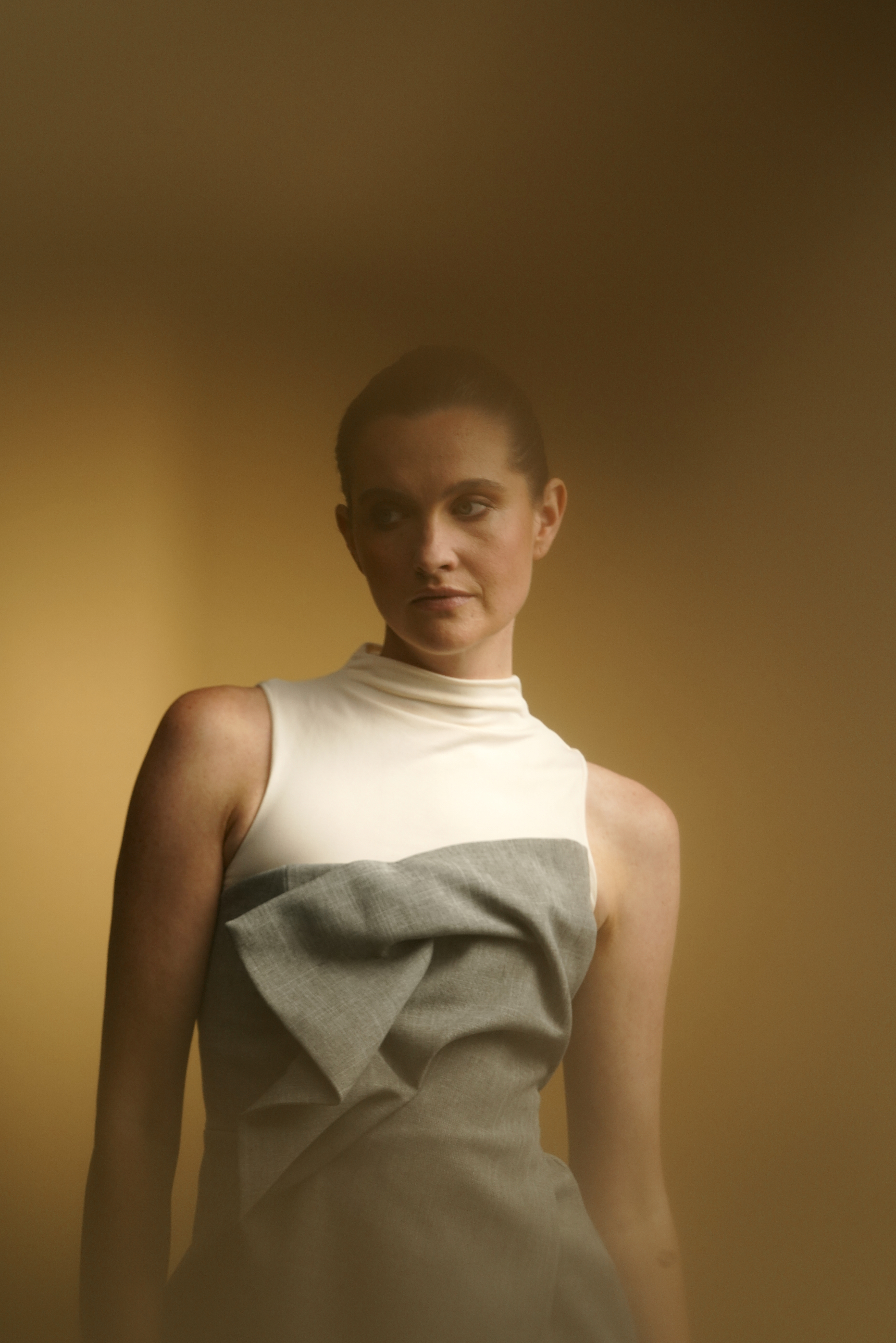 Een vrouw met een mouwloze jurk met hoge kraag en strikdetail staat voor een onscherpe achtergrond in warme tinten.