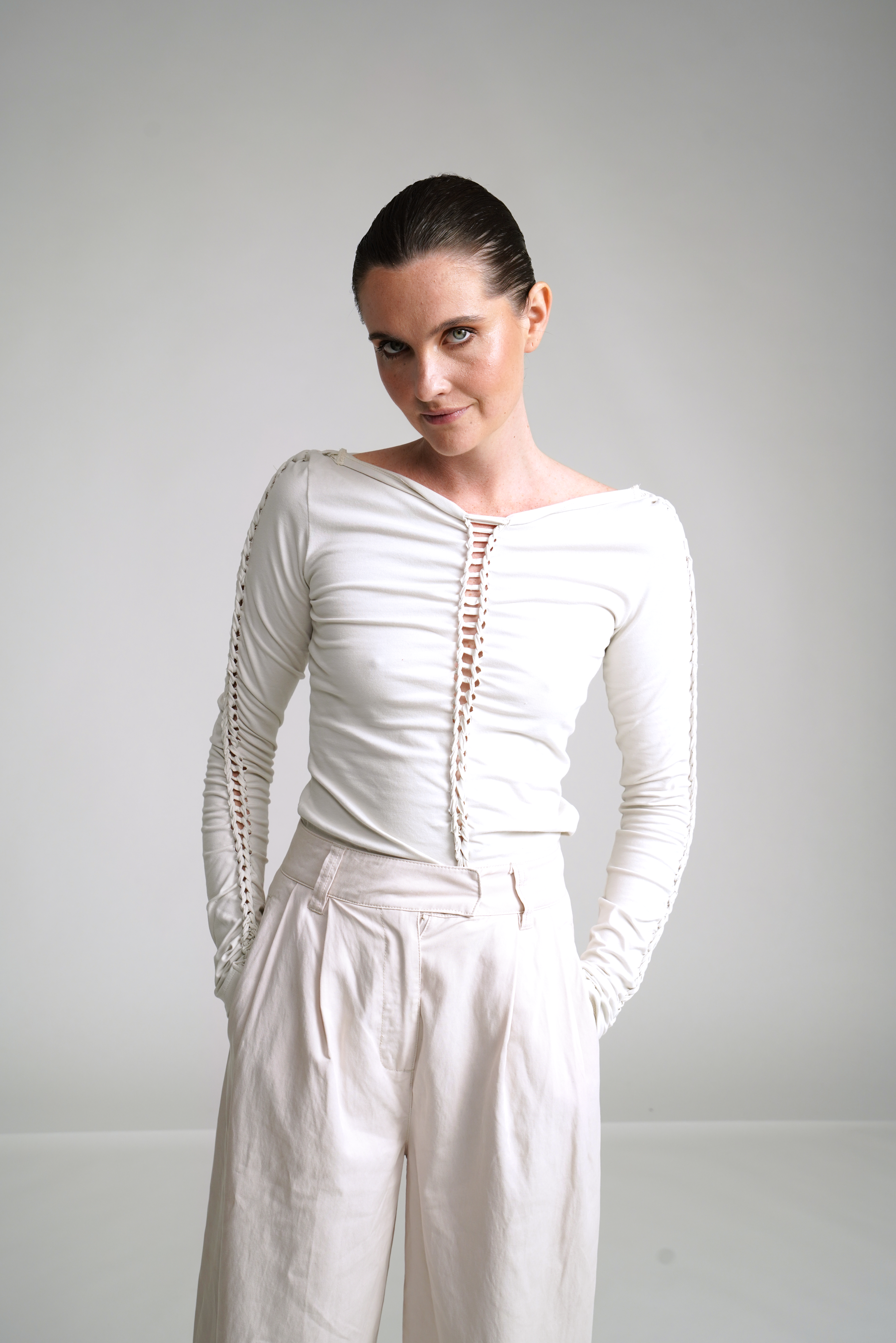 A person in a white, long-sleeved top with a unique front design and light pants stands against a plain background, hands in pockets.