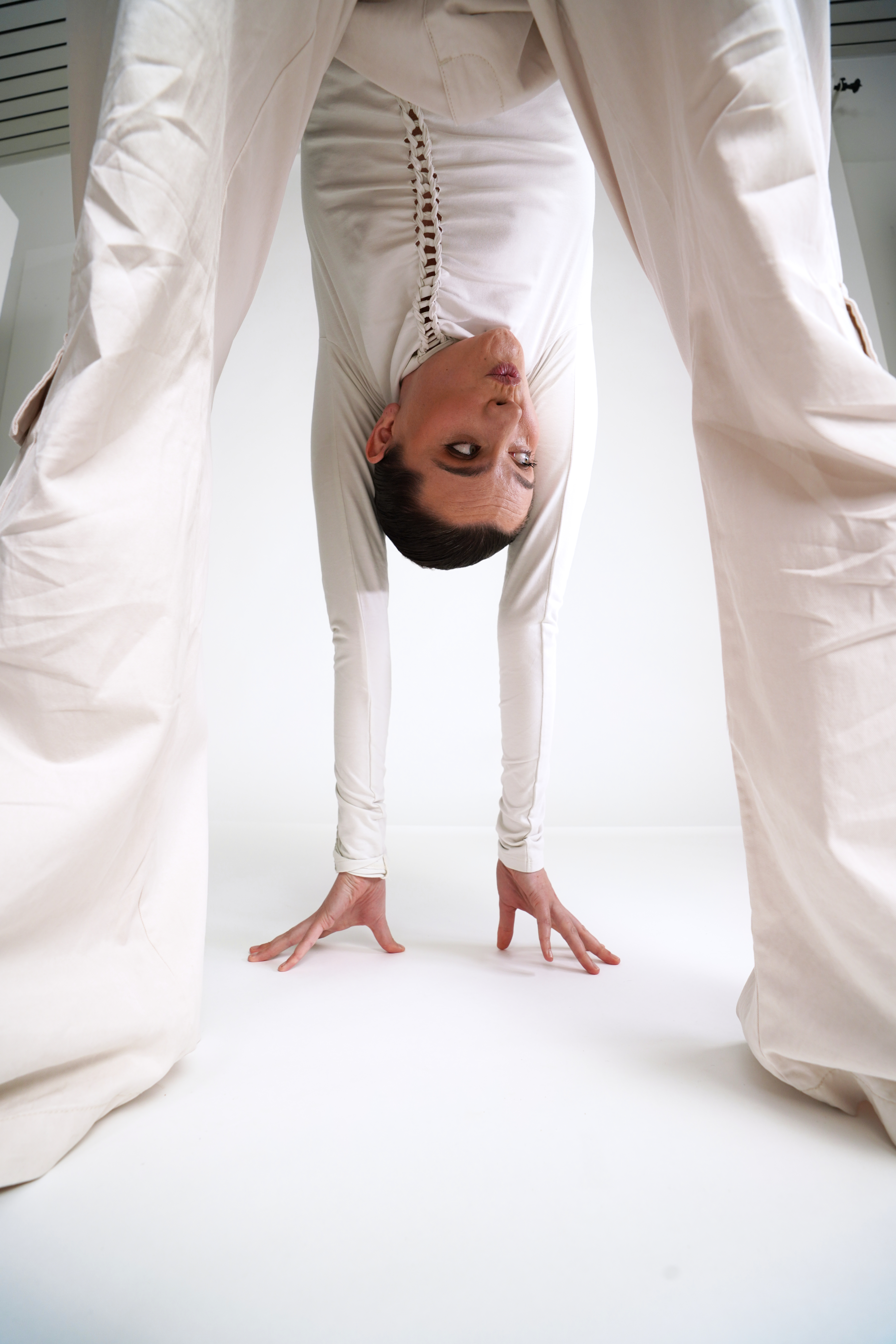 Person in white clothing bending over, looking through legs with hands on the floor, in a minimalist white room.