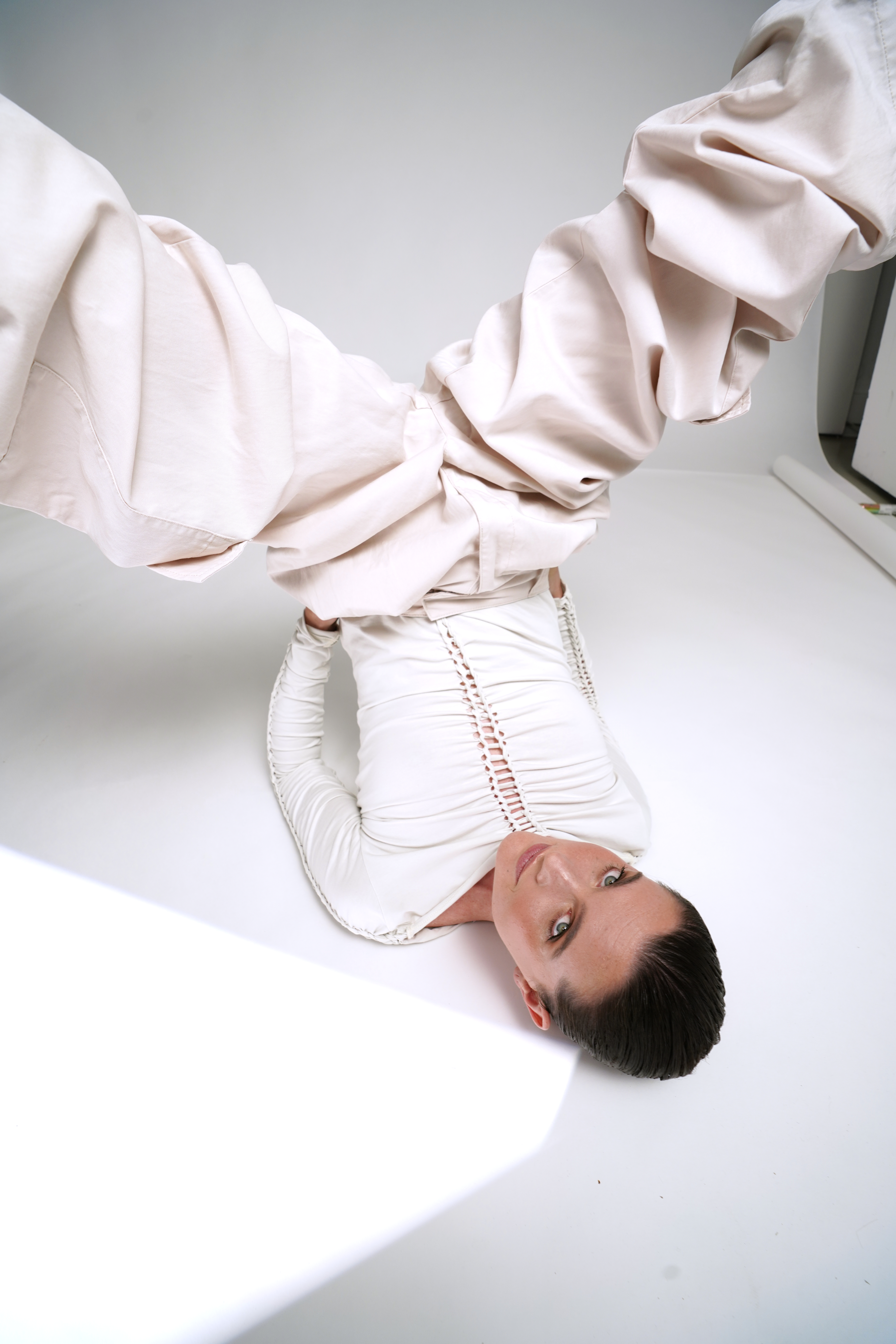 Person in a white outfit with voluminous pants poses upside down on a white floor, looking up with a neutral expression.