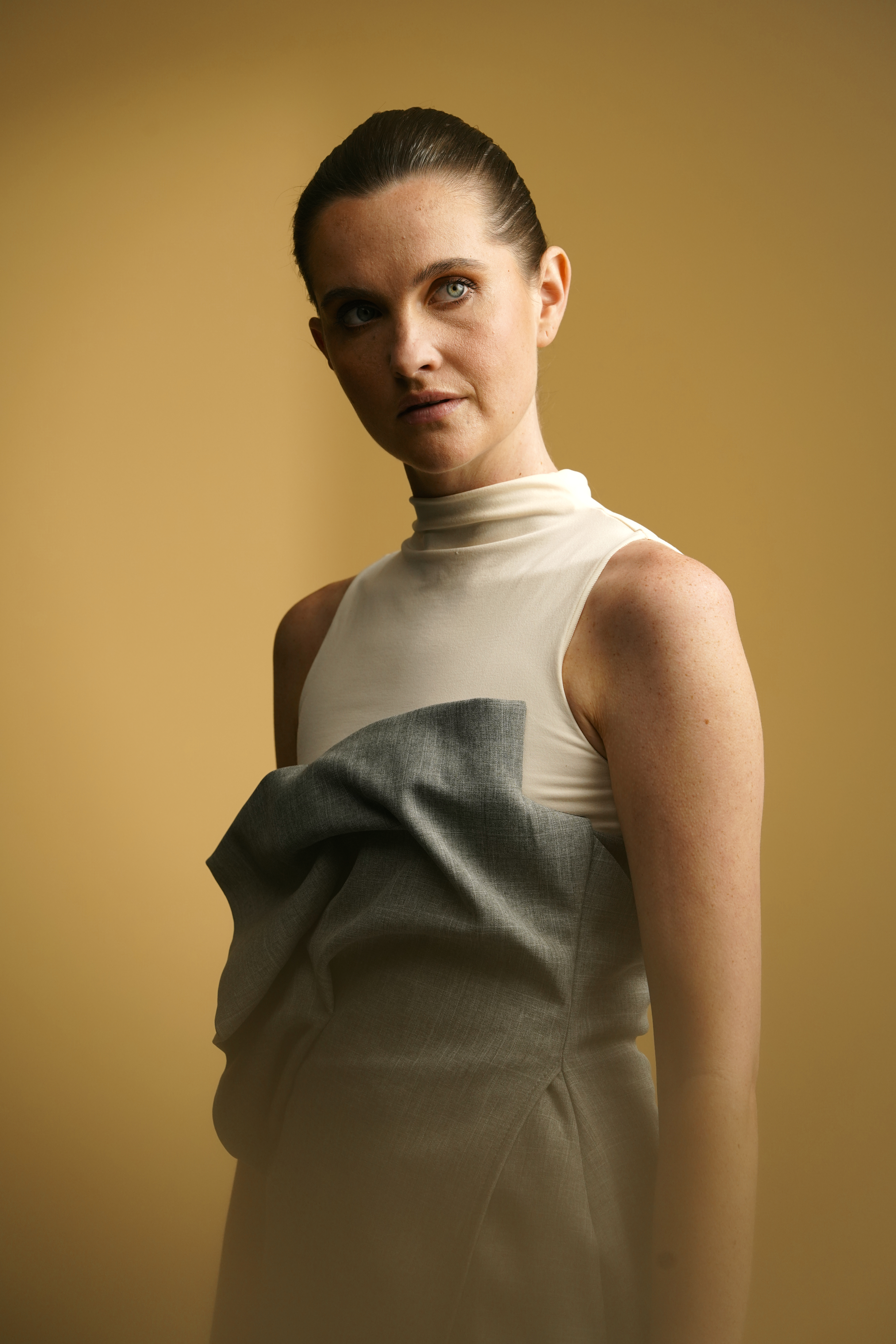 Een vrouw met een wit mouwloos topje en een grijze jurk poseert voor een beige achtergrond en kijkt lichtjes opzij.