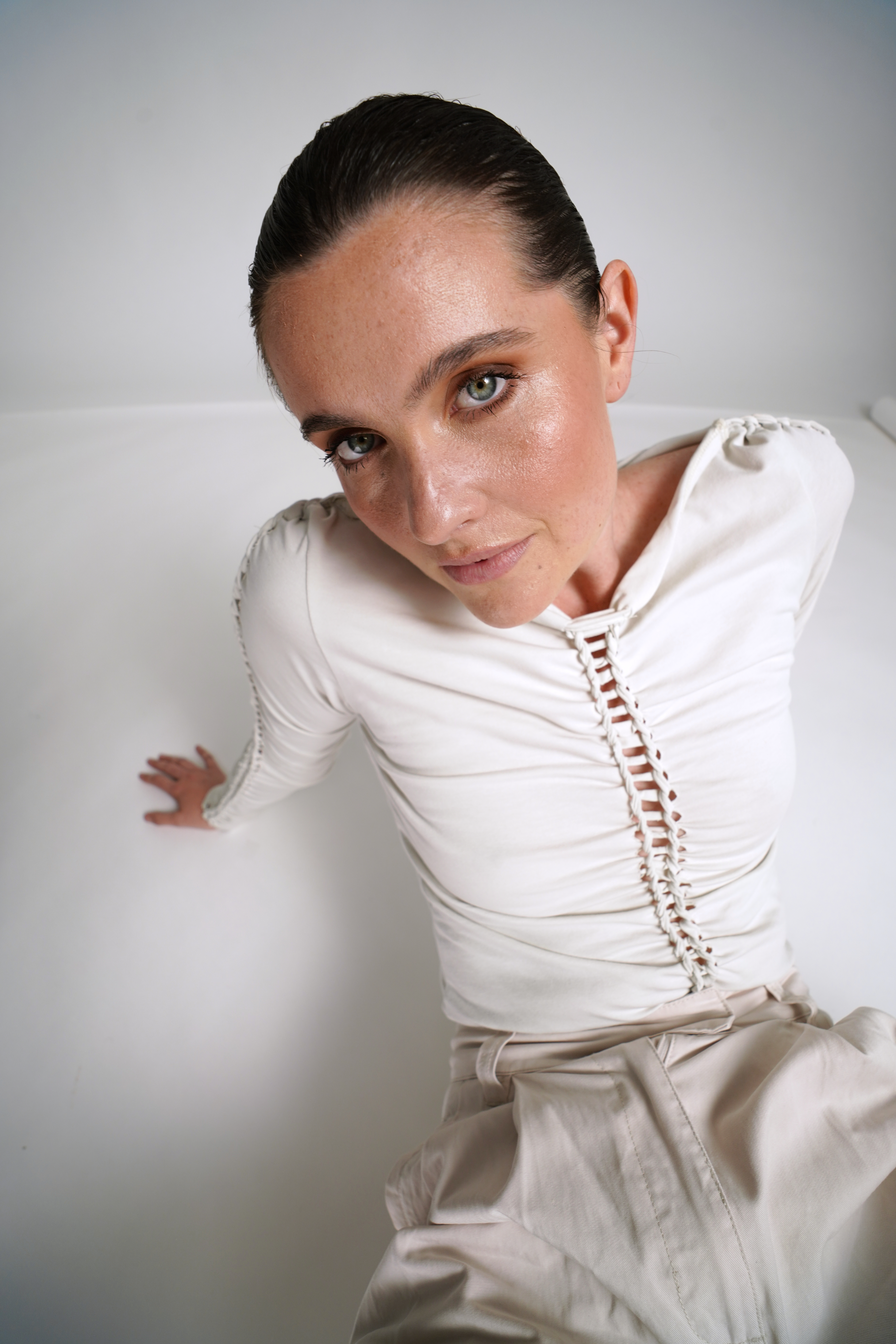 Woman in a white, ruched top and light pants, sitting on the floor, looking up at the camera with a neutral expression.