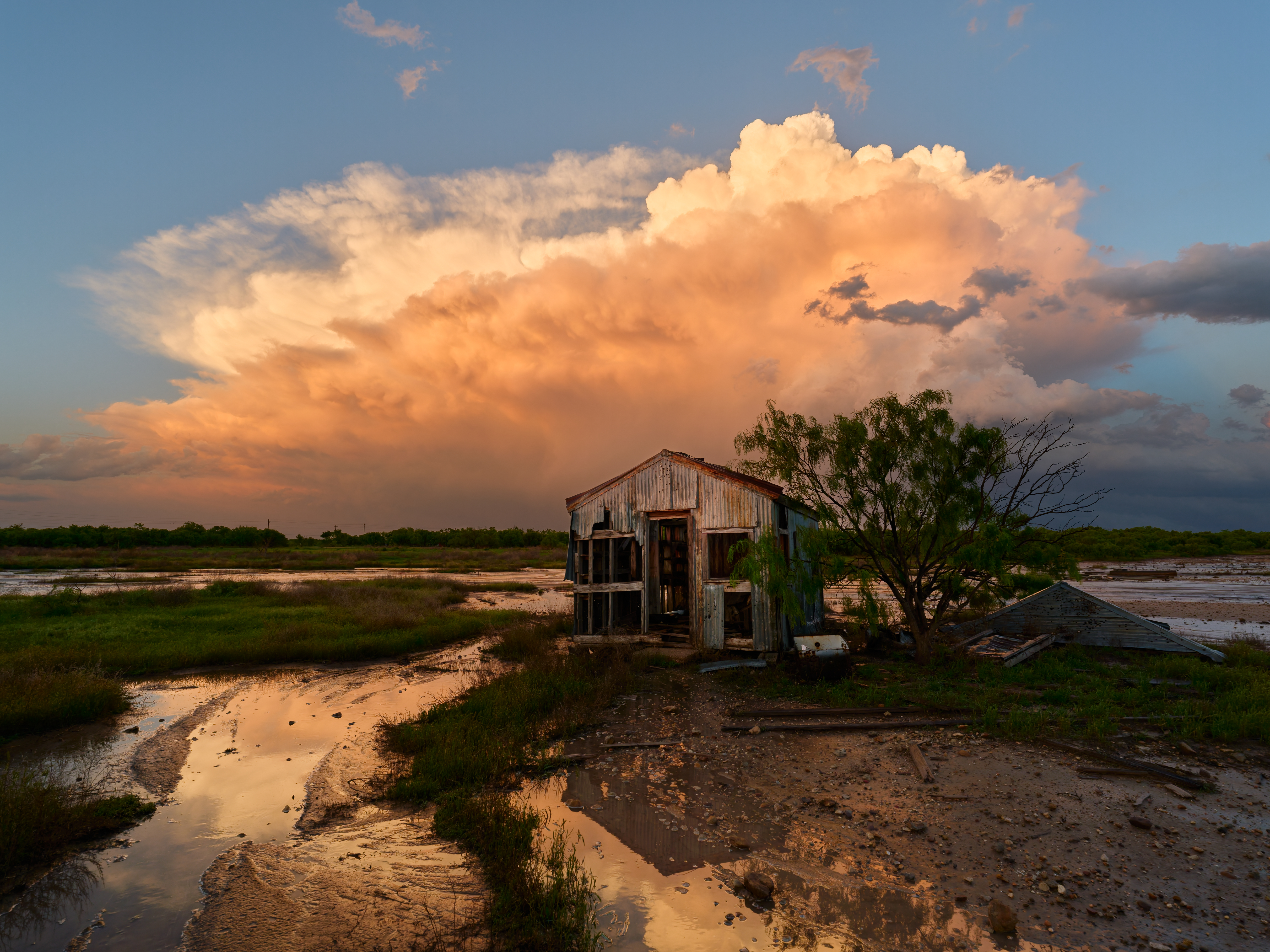 A weathered shack stands in a flooded field at sunset, with vibrant clouds and a tree nearby, reflecting in the water.