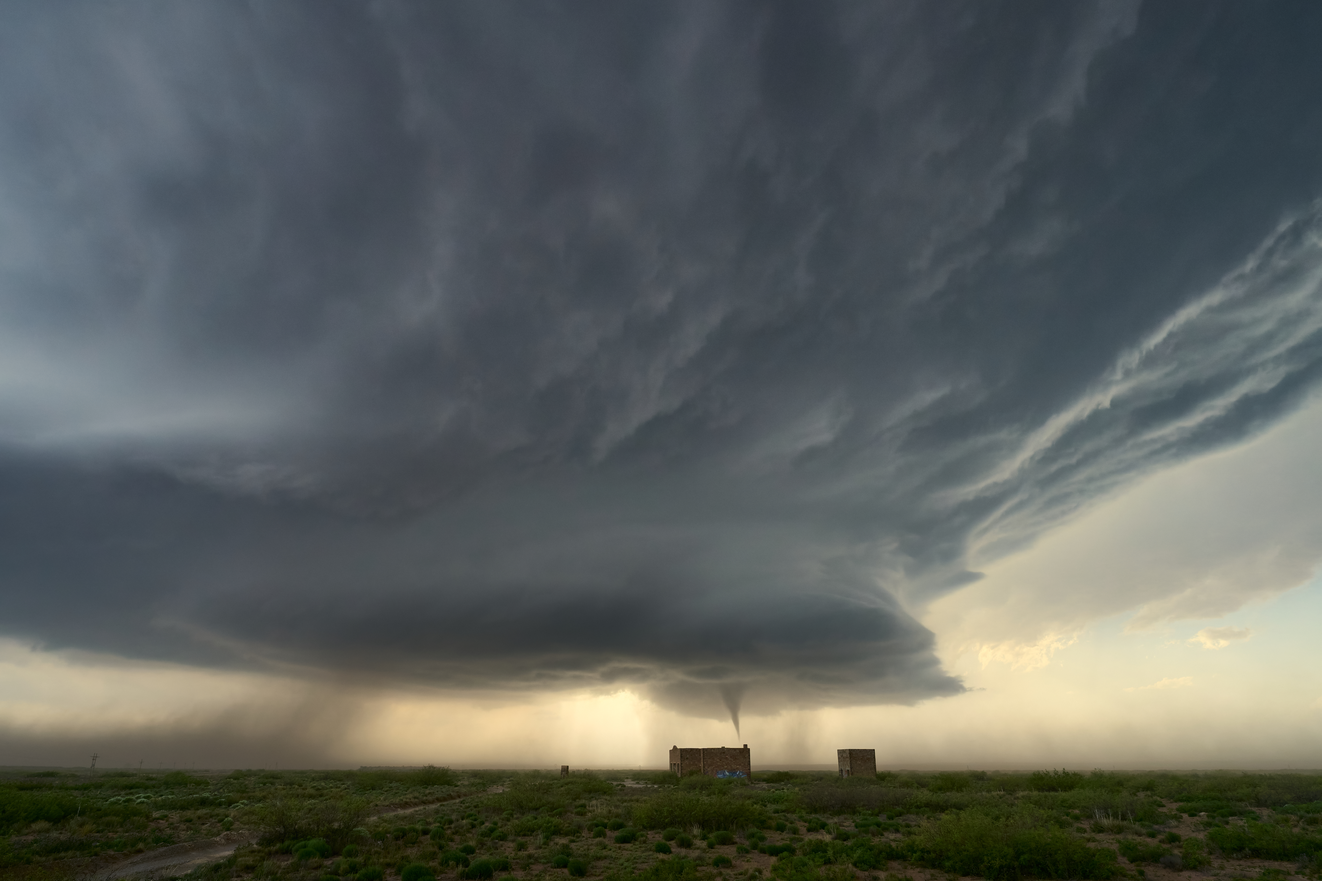 A vast stormy sky with dark clouds looms over a small house on a flat landscape, with a visible tornado forming in the distance.