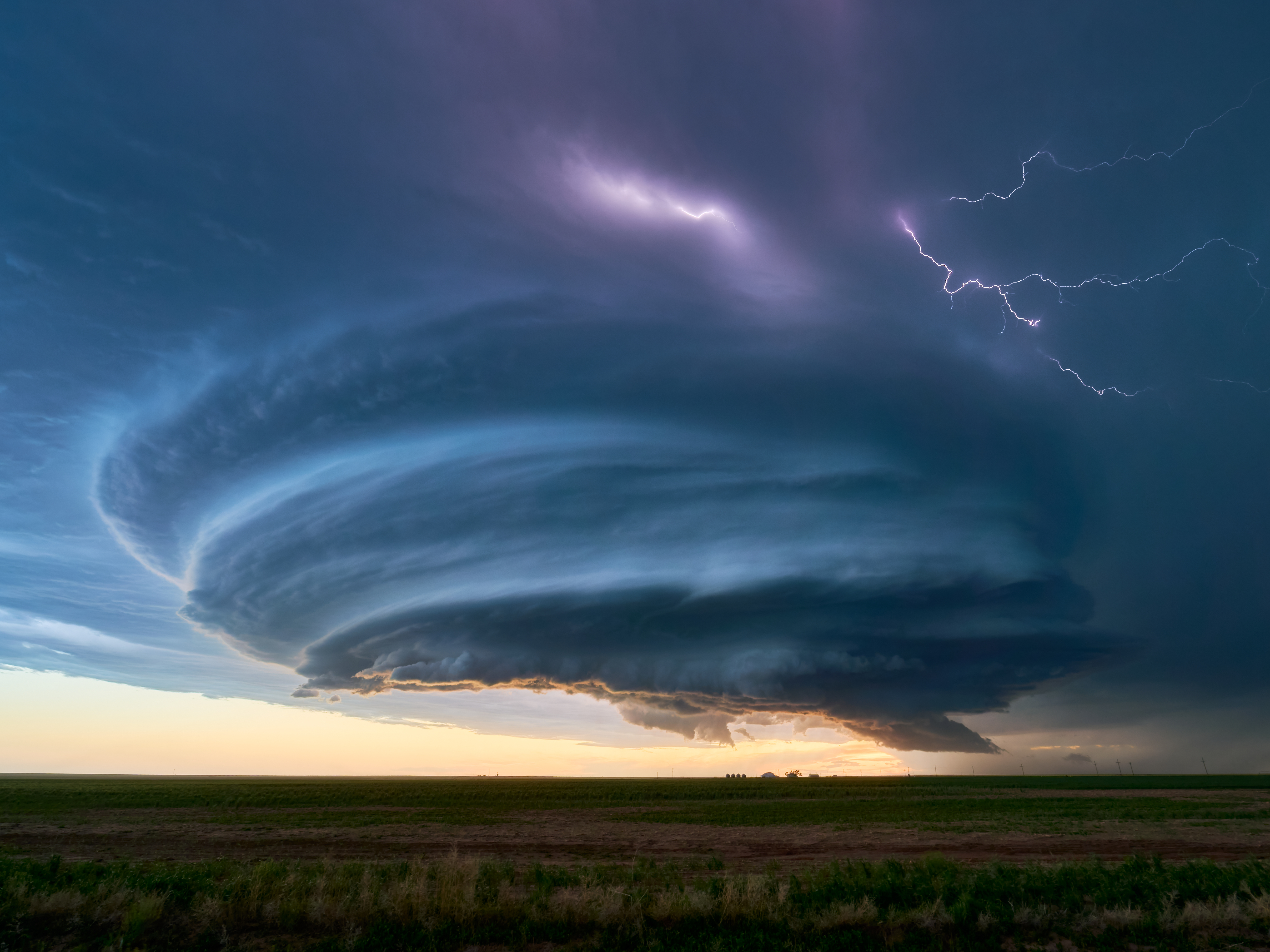 A massive, swirling supercell thunderstorm with lightning illuminates the sky over a flat landscape at sunset.