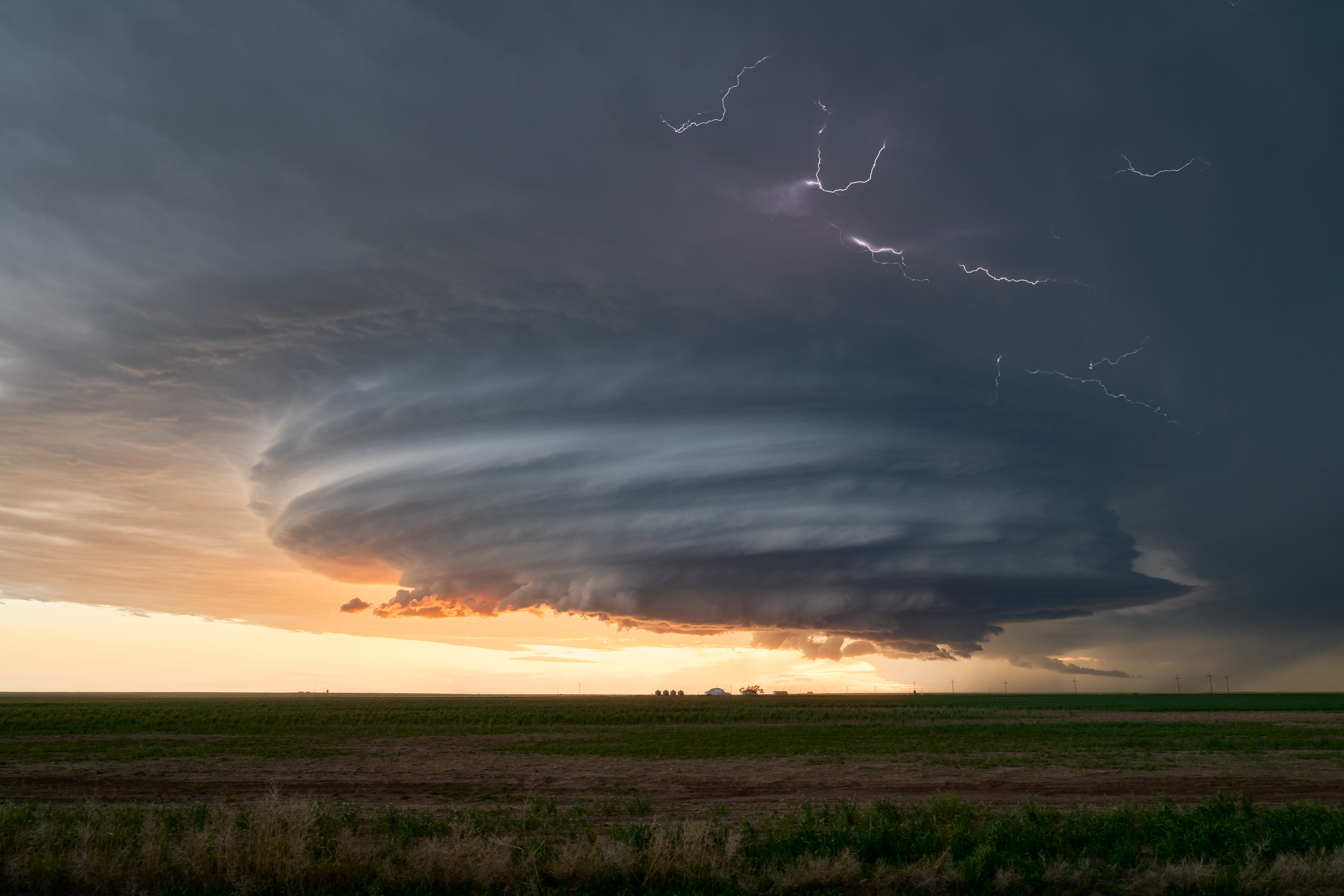 A dramatic supercell thunderstorm with swirling clouds and lightning over a flat landscape at sunset.
