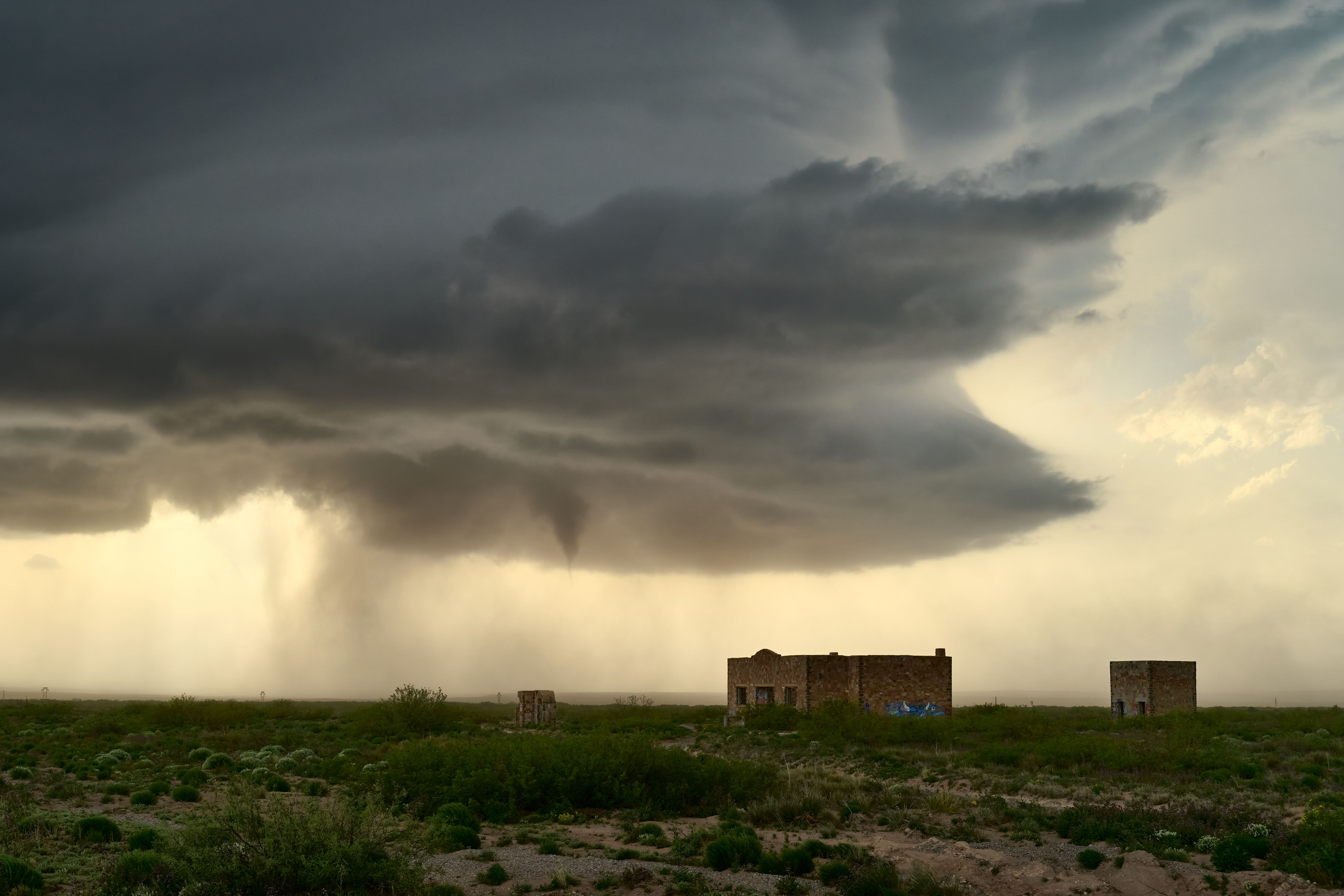 A dark, swirling storm cloud with a funnel shape looms over a grassy field and two small, abandoned buildings.