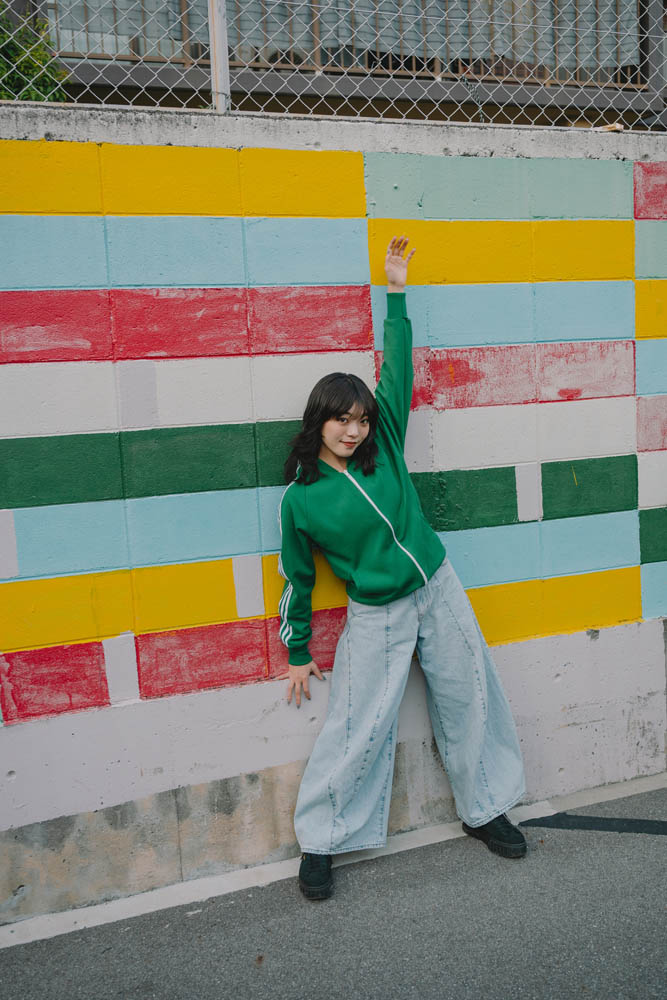 A person in a green jacket and wide blue jeans poses cheerfully against a colorful tiled wall with red, yellow, and light blue blocks.