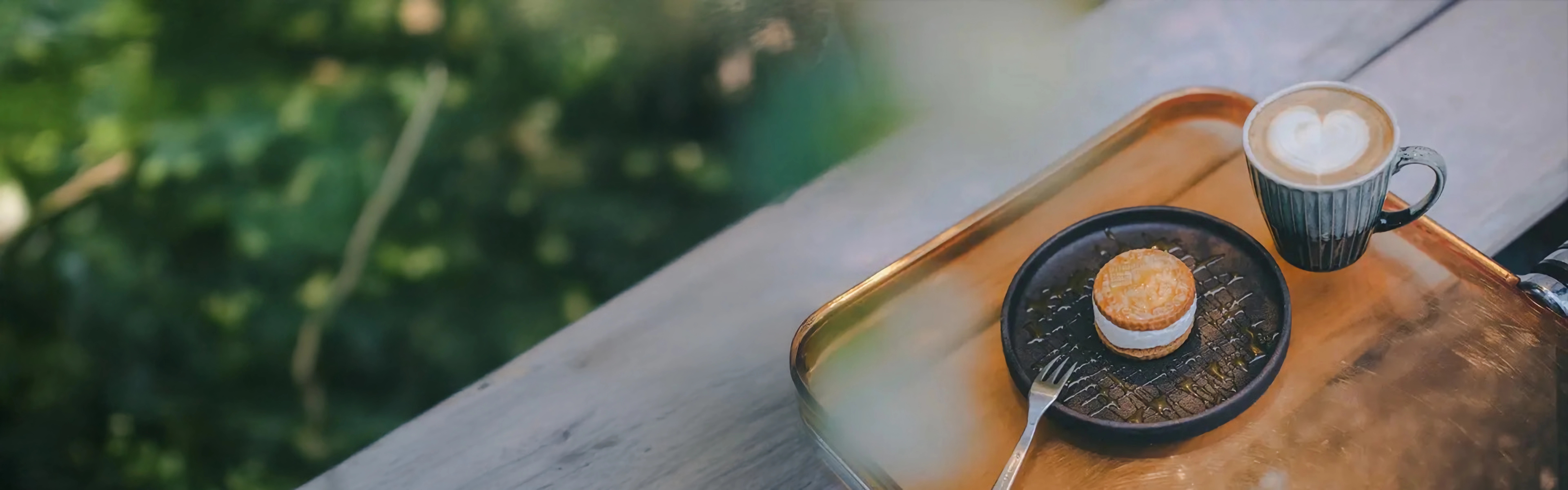 Ein Holztisch mit einem Tablett, darauf eine Tasse Kaffee und ein kleiner Kuchen auf einem Teller, vor einem unscharfen, grünen Naturhintergrund.