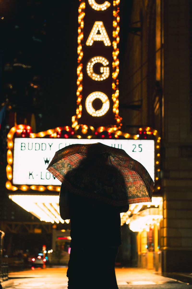 Silhouette of a person with an umbrella in front of a neon-lit theater sign that reads "CAGO" at night.