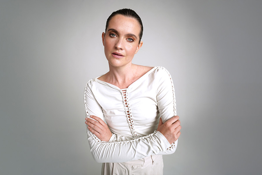 Person with slicked-back hair in a white top, arms crossed, against a plain gray background.