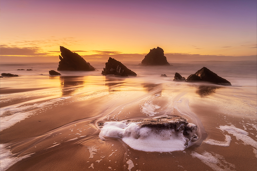 Sonnenuntergang über einem ruhigen Strand, an dem sich schäumende Wellen an den Felsen brechen und orangefarbene und violette Farbtöne am Himmel und im nassen Sand widerspiegeln.