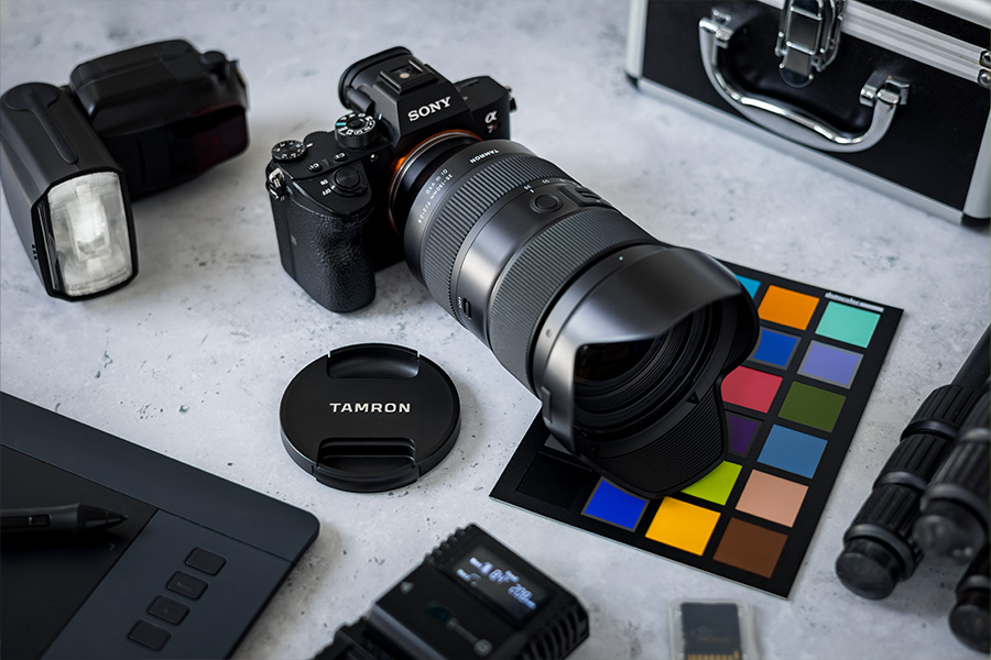 Kameraausrüstung auf einem Tisch: Eine DSLR mit Objektiv, Objektivdeckel, Blitz, Colorchecker und ein Tablet – alles auf einer hellgrauen Oberfläche arrangiert.
