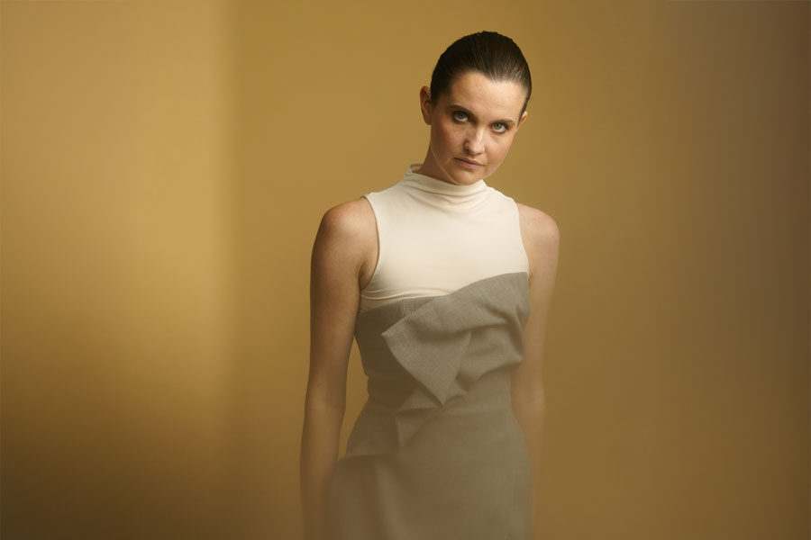 A woman in a sleeveless dress with a high collar stands against a warm, golden background, looking directly at the camera.