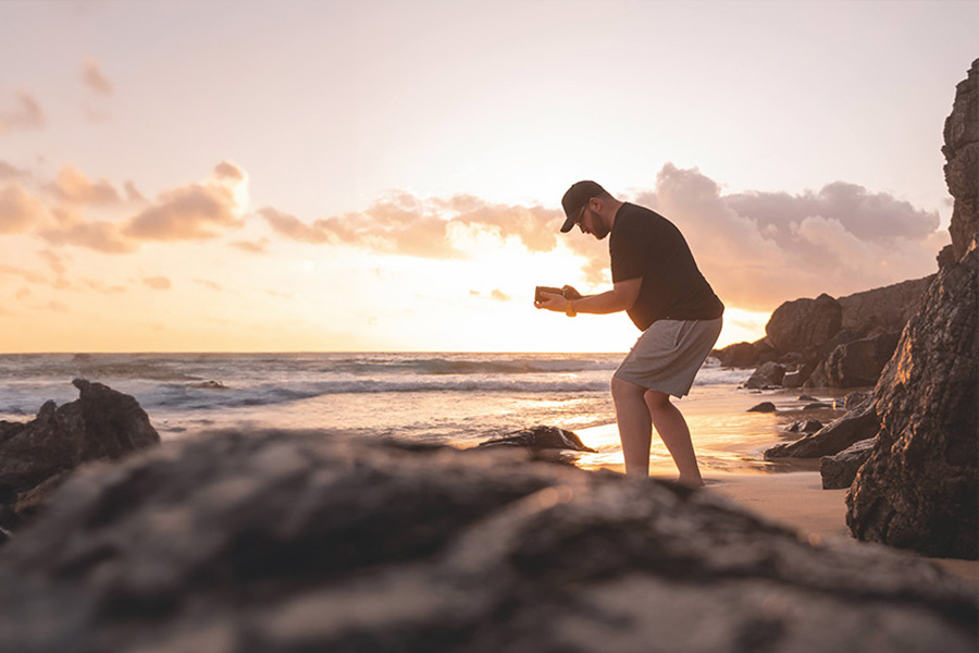 Una persona è in piedi su una spiaggia rocciosa al tramonto, mentre scatta foto con una fotocamera. Il cielo è coperto di nuvole e le onde si infrangono sulla riva.