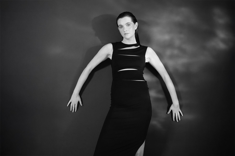 Eine Frau im schwarzen Kleid posiert vor einem dunklen Hintergrund. Das Licht wirft Schatten an die Wand und verleiht der minimalistischen Komposition Tiefe.