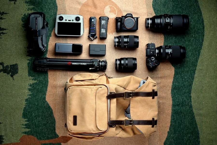 Equipo fotográfico extendido sobre una superficie estampada, incluyendo cámaras, objetivos, un trípode y una bolsa de cámara de color marrón claro.