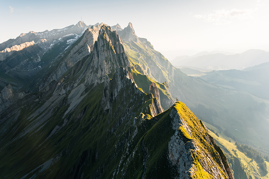 Bergrücken im flachen Abendlicht mit scharfen Gipfeln, bedeckt mit Gras und Felsen, unter einem klaren Himmel, mit Panoramablick auf ferne Täler.