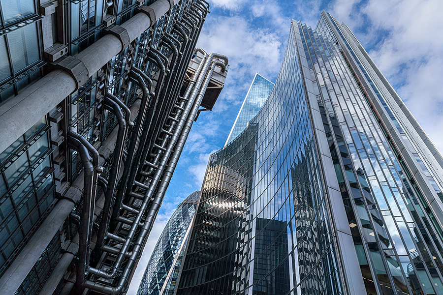 Skyscrapers with reflective glass and industrial pipes under a cloudy sky, creating a modern cityscape.
