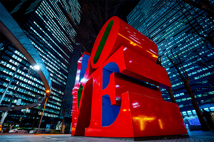 Una scultura rossa e blu con la scritta «LOVE» si erge in un paesaggio urbano notturno, circondata da alti edifici illuminati.