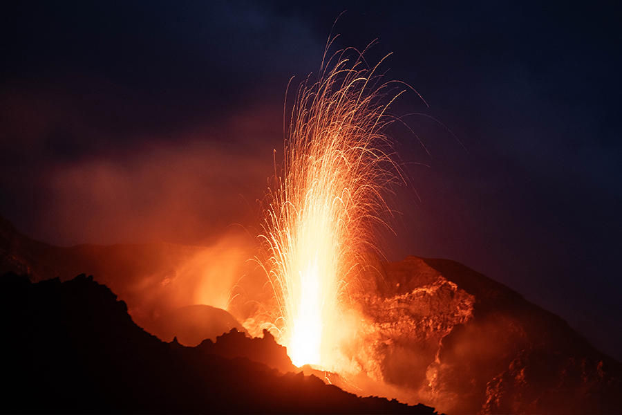 Ein Vulkan, der nachts ausbricht, spuckt hellorange Lava und Funken in den dunklen Himmel, während dichter Rauch aus dem Krater aufsteigt.