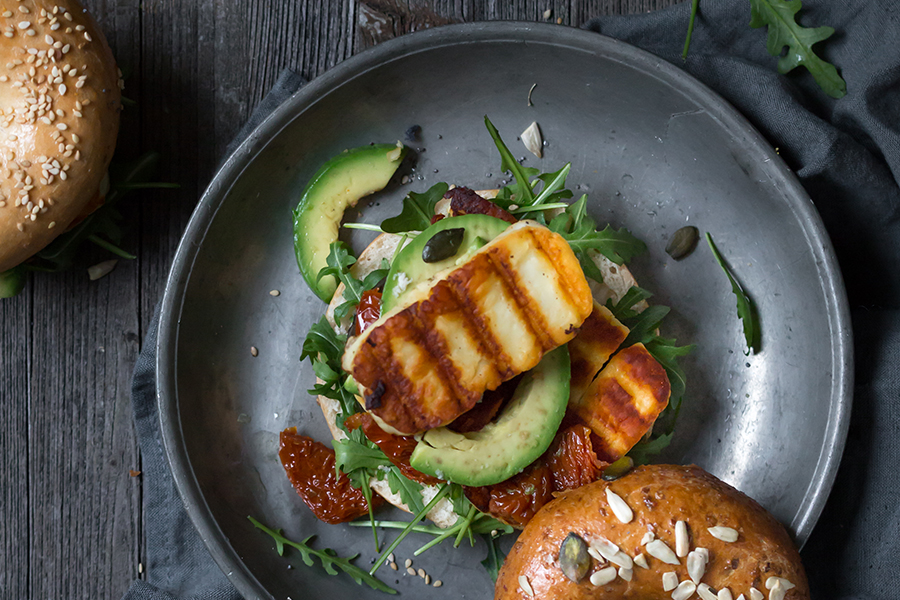 Gegrillter Halloumi, Avocado, sonnengetrocknete Tomaten und Rucola auf einem Teller, dazu Körnerbrötchen auf einem rustikalen Holztisch.